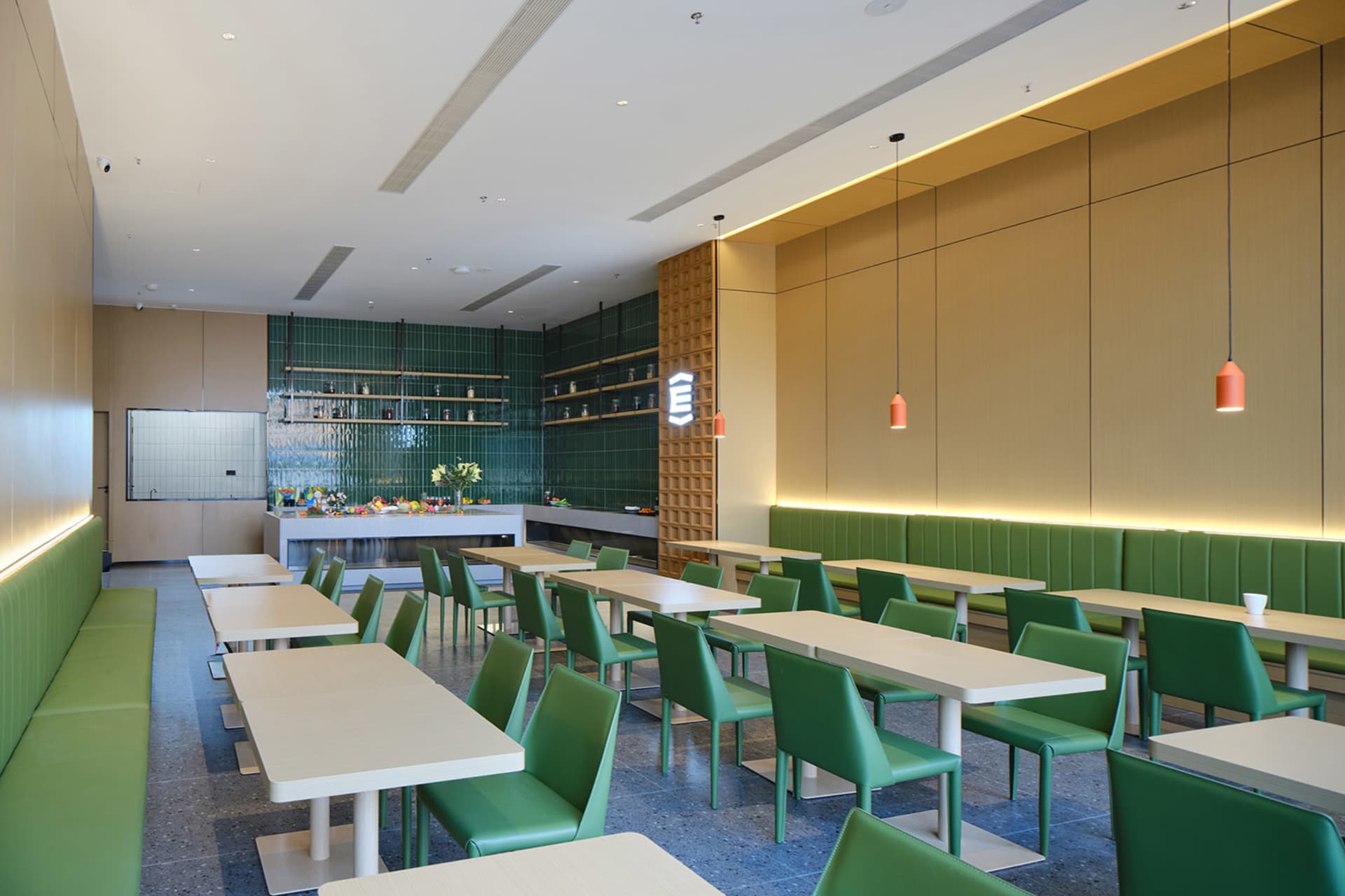 Modern cafeteria interior with rows of white tables, green leather chairs and booths, green tiled feature wall, and warm wood paneling under pendant lighting.