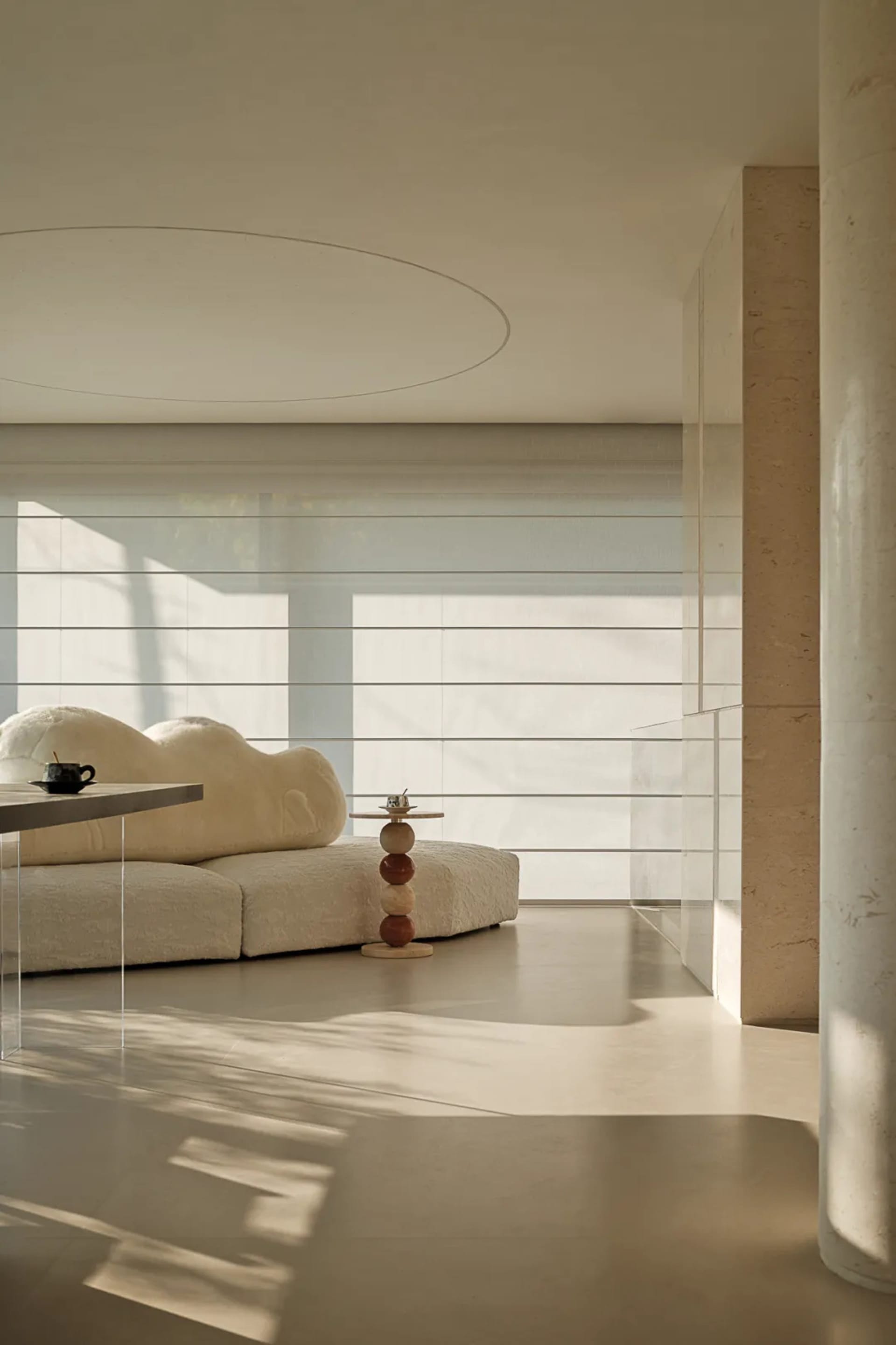 Minimalist living room featuring a white cloud-shaped modular sofa, travertine walls and columns, acrylic side table, and a sculptural wooden side table under a circular ceiling detail.