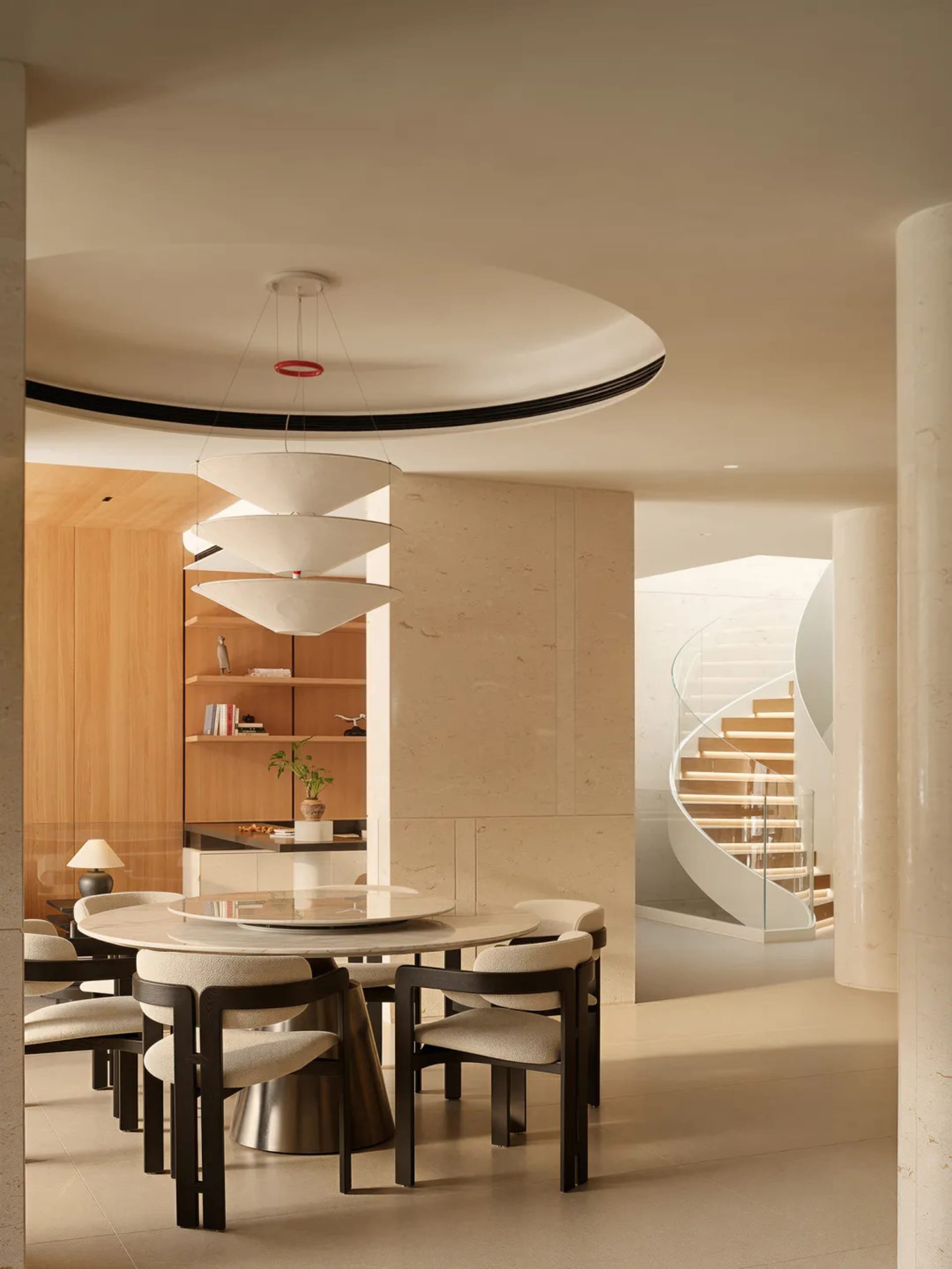 Modern dining room featuring a round marble table, dark wood upholstered chairs, a large multi-tiered pendant light, travertine walls, and a dramatic white spiral staircase in the background.