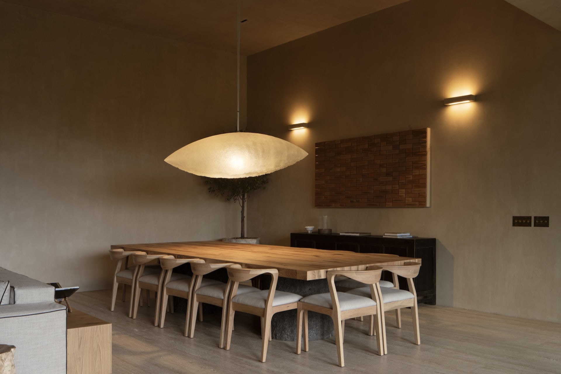 Modern minimalist dining room featuring a long solid oak dining table, eight matching oak dining chairs with light upholstery, a large sculptural pendant lamp, dark wood sideboard, and textured plaster walls in warm neutral tones.