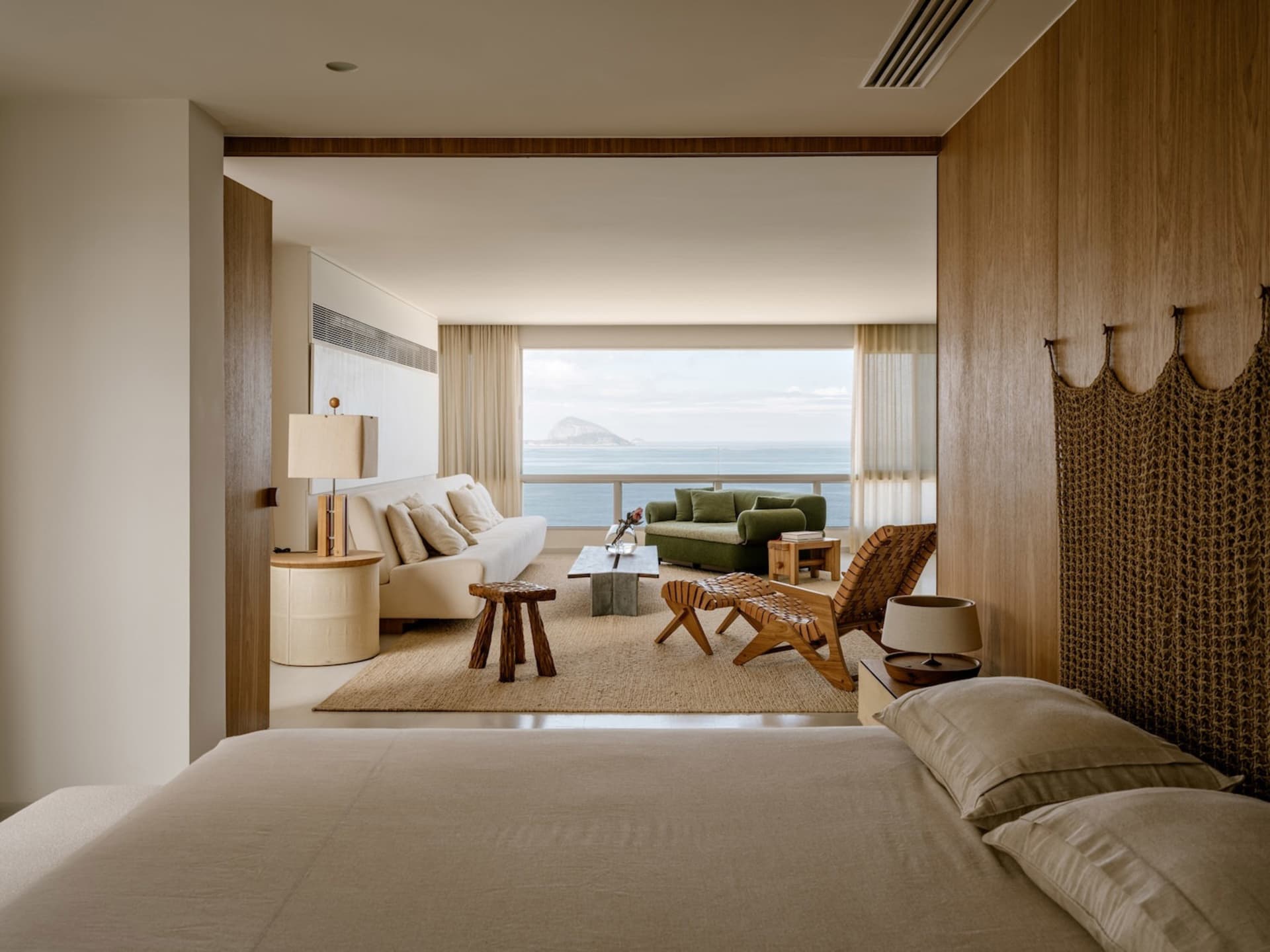 Modern coastal bedroom with beige linen bedding, woven teak headboard, and panoramic ocean view through the adjacent living room.