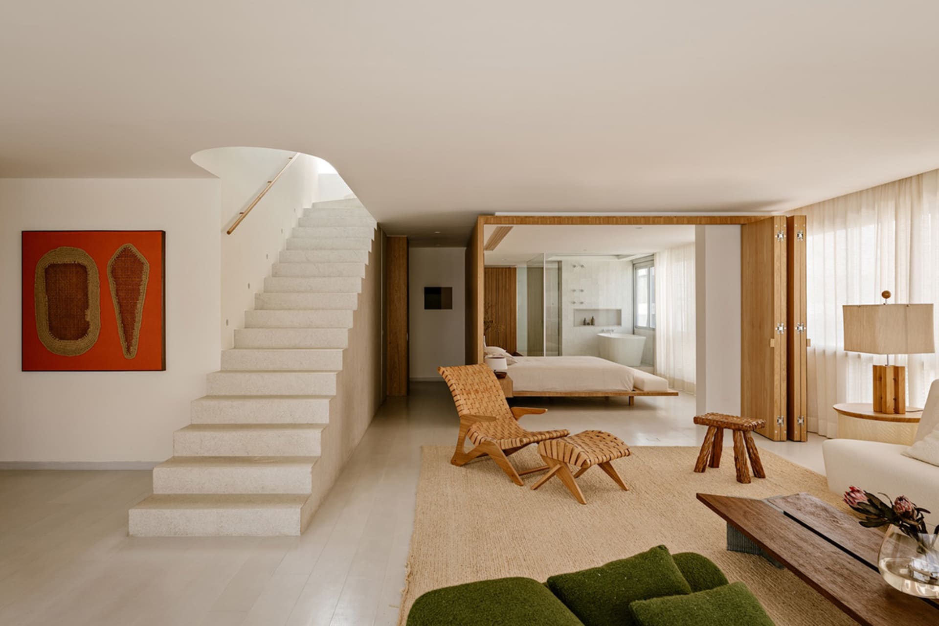 Modern open-plan living room featuring a woven rattan lounge chair, green bouclé sofa, stone staircase, and view into a bedroom with freestanding bathtub.