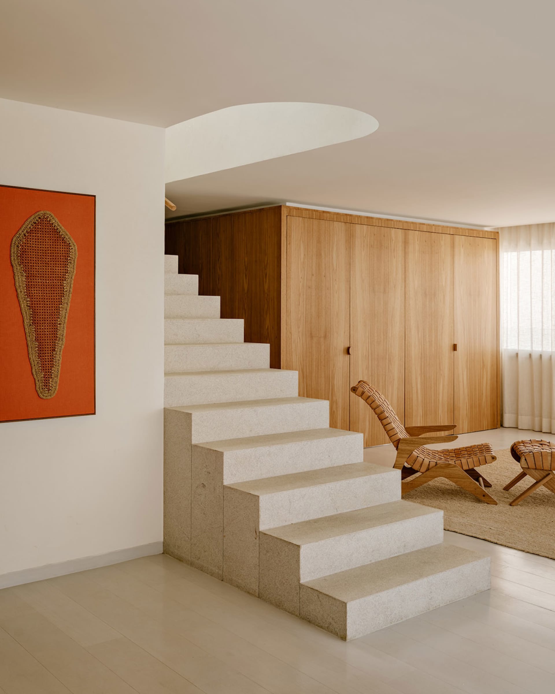 Modern interior featuring a floating terrazzo staircase, light wood built-in storage, woven rattan lounge chair and ottoman, and an orange artwork with woven textile on a white wall.