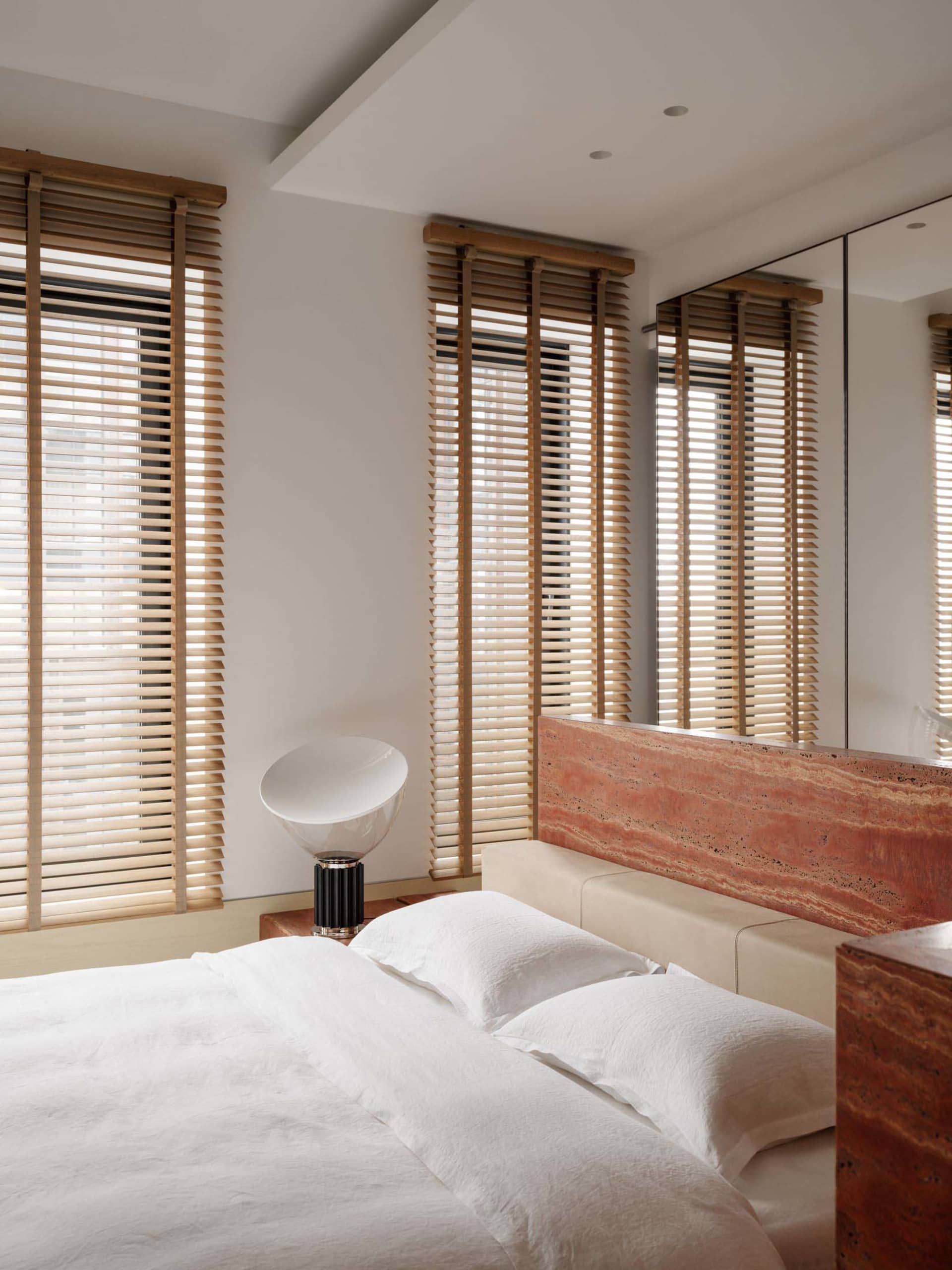 Modern minimalist bedroom with a large red travertine stone headboard, white bedding, wooden Venetian blinds, and a sculptural white table lamp.