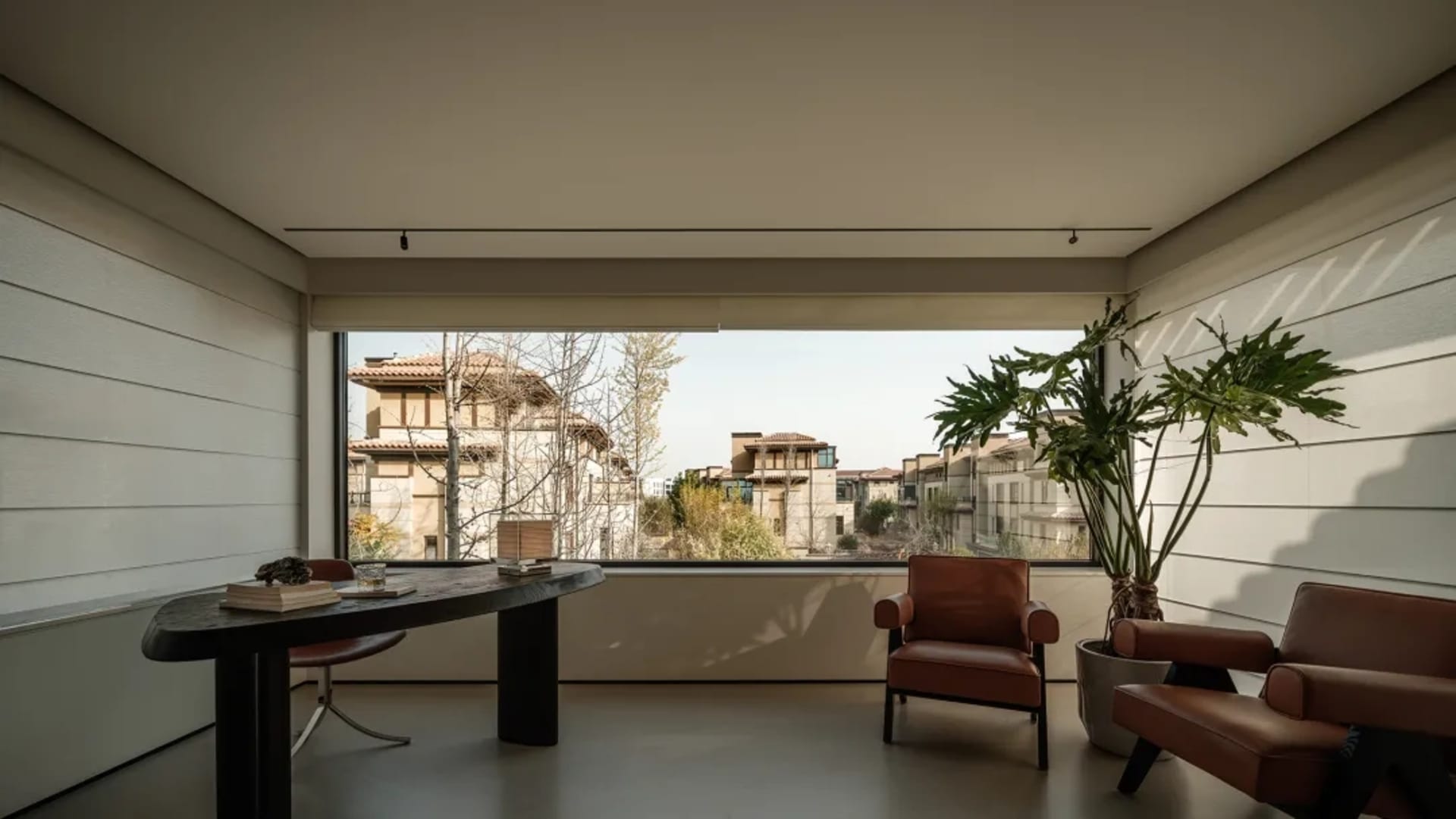 Modern minimalist home office featuring two cognac leather armchairs, a dark wood oval desk, large picture window with city view, and a tall potted plant.