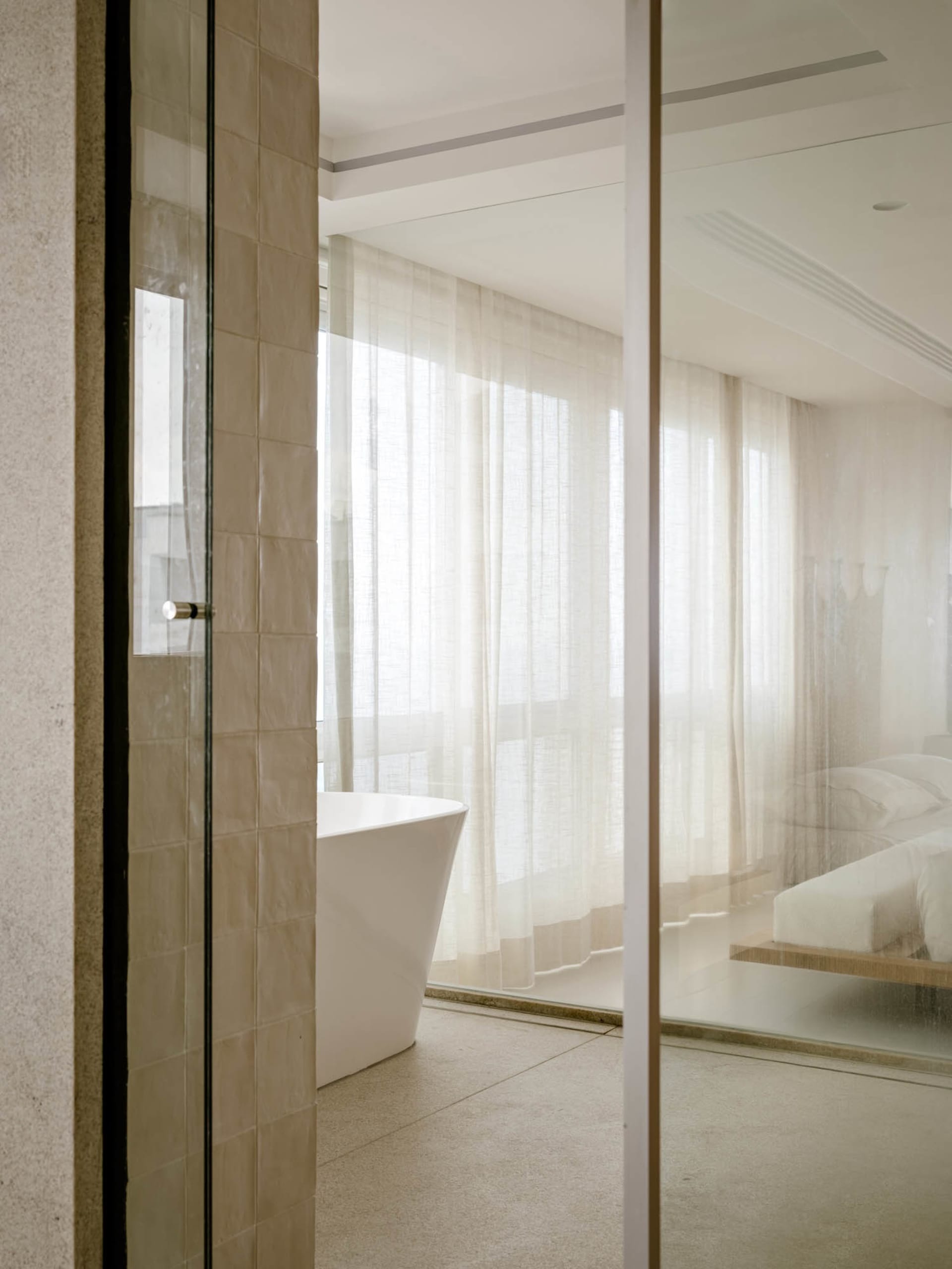 Modern minimalist bathroom interior with beige travertine tiled walls, white freestanding bathtub, sheer white curtains, and a floating bed visible through glass partition.