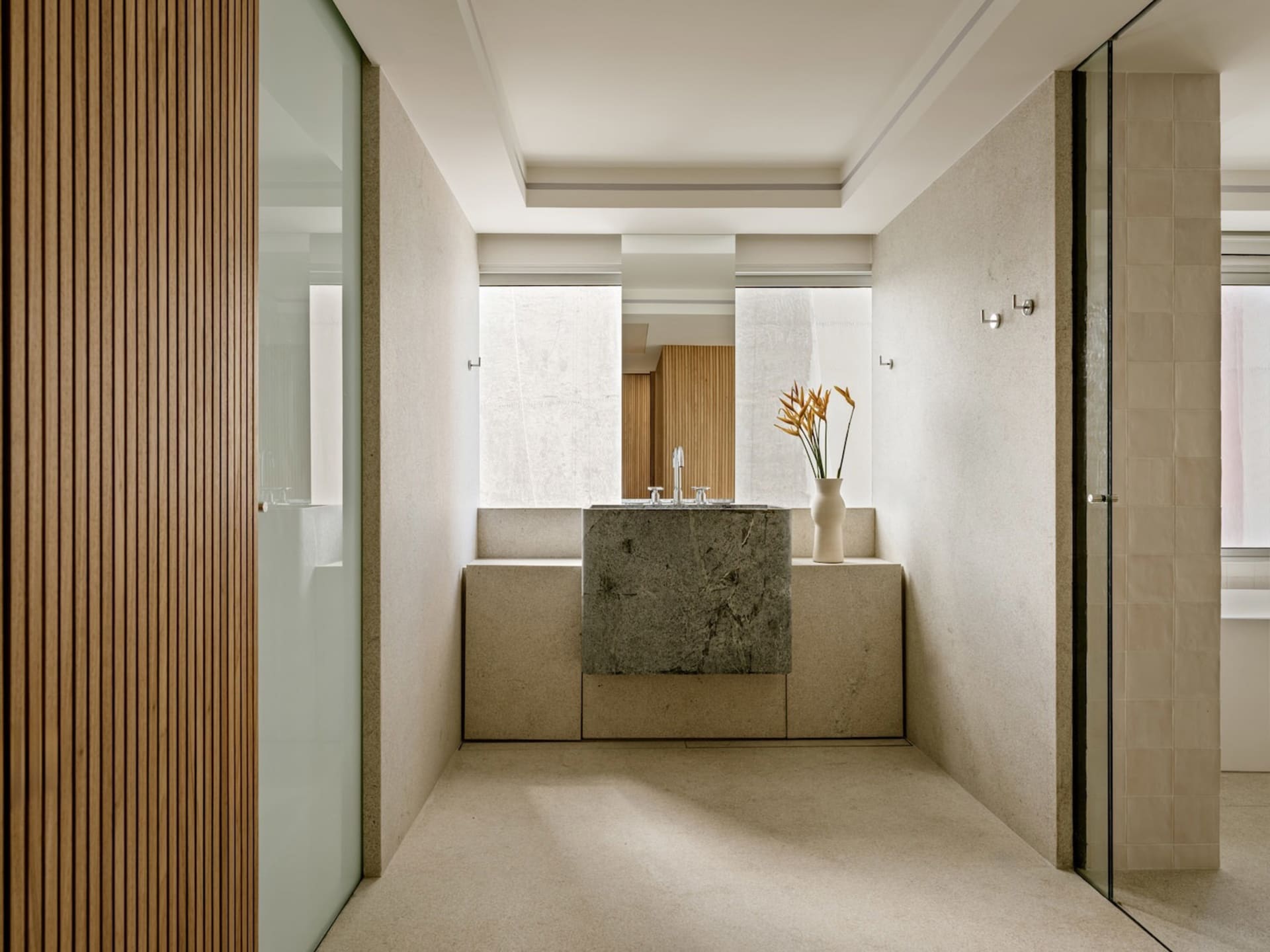 Modern minimalist bathroom with a large monolithic dark stone vanity, travertine walls and floor, vertical wood slat accent wall, and a vase of orange flowers.