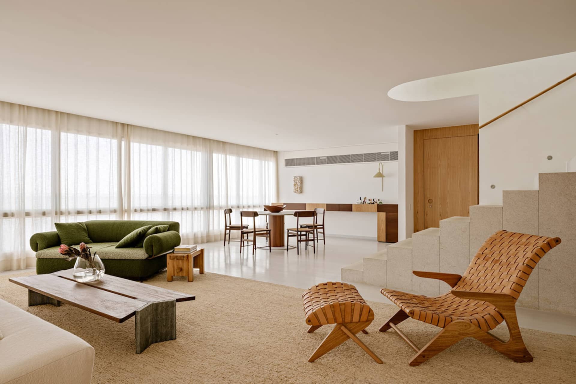 Modern open-plan living room featuring a moss-green velvet sofa, a sculptural woven leather lounge chair and ottoman, a live-edge wooden coffee table, and a dining area in the background with light wood cabinetry and sheer curtains.