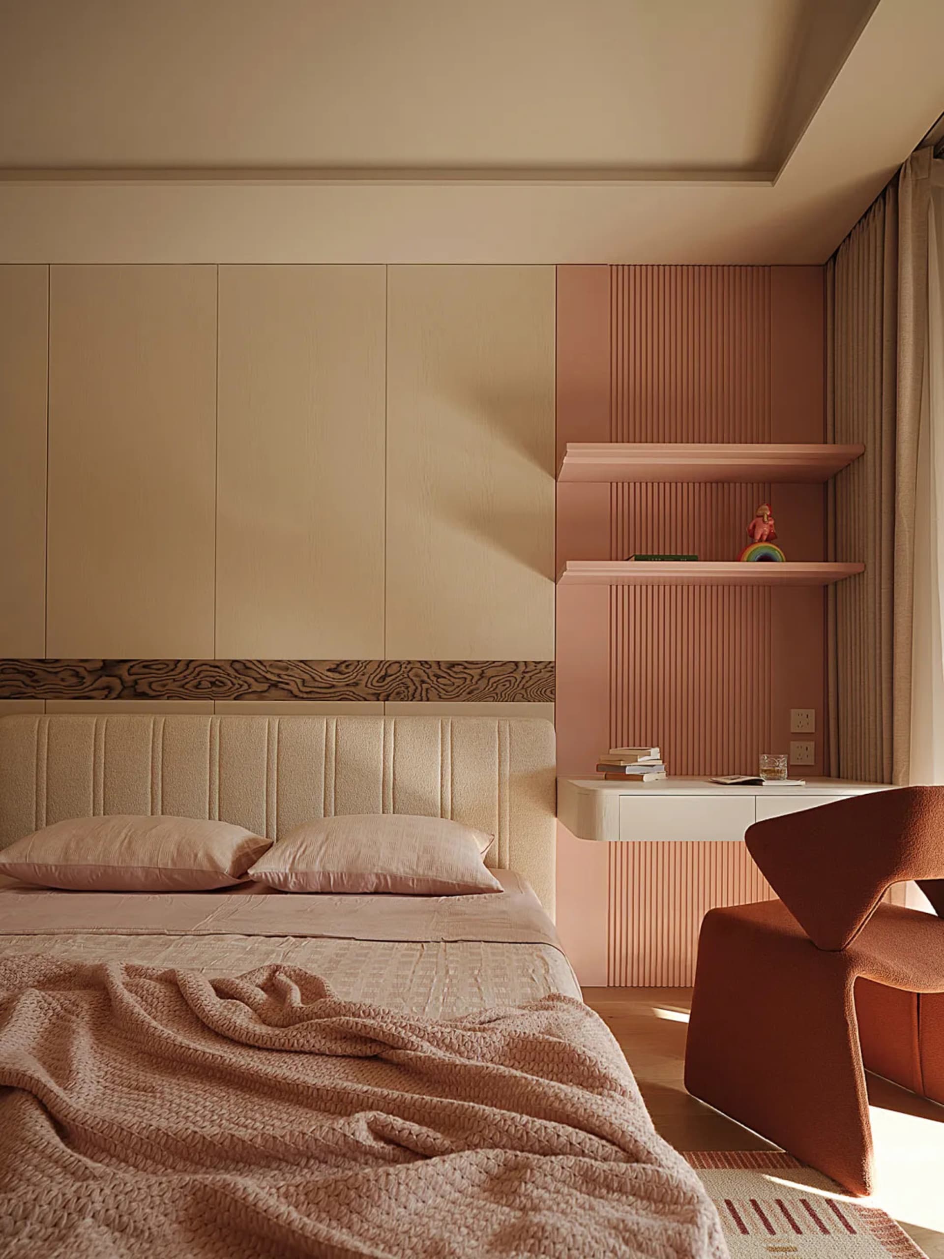 Modern bedroom featuring a beige upholstered bed with pink bedding, a terracotta sculptural armchair, and a two-tone wall with beige cabinetry and a pink fluted accent wall with floating shelves.