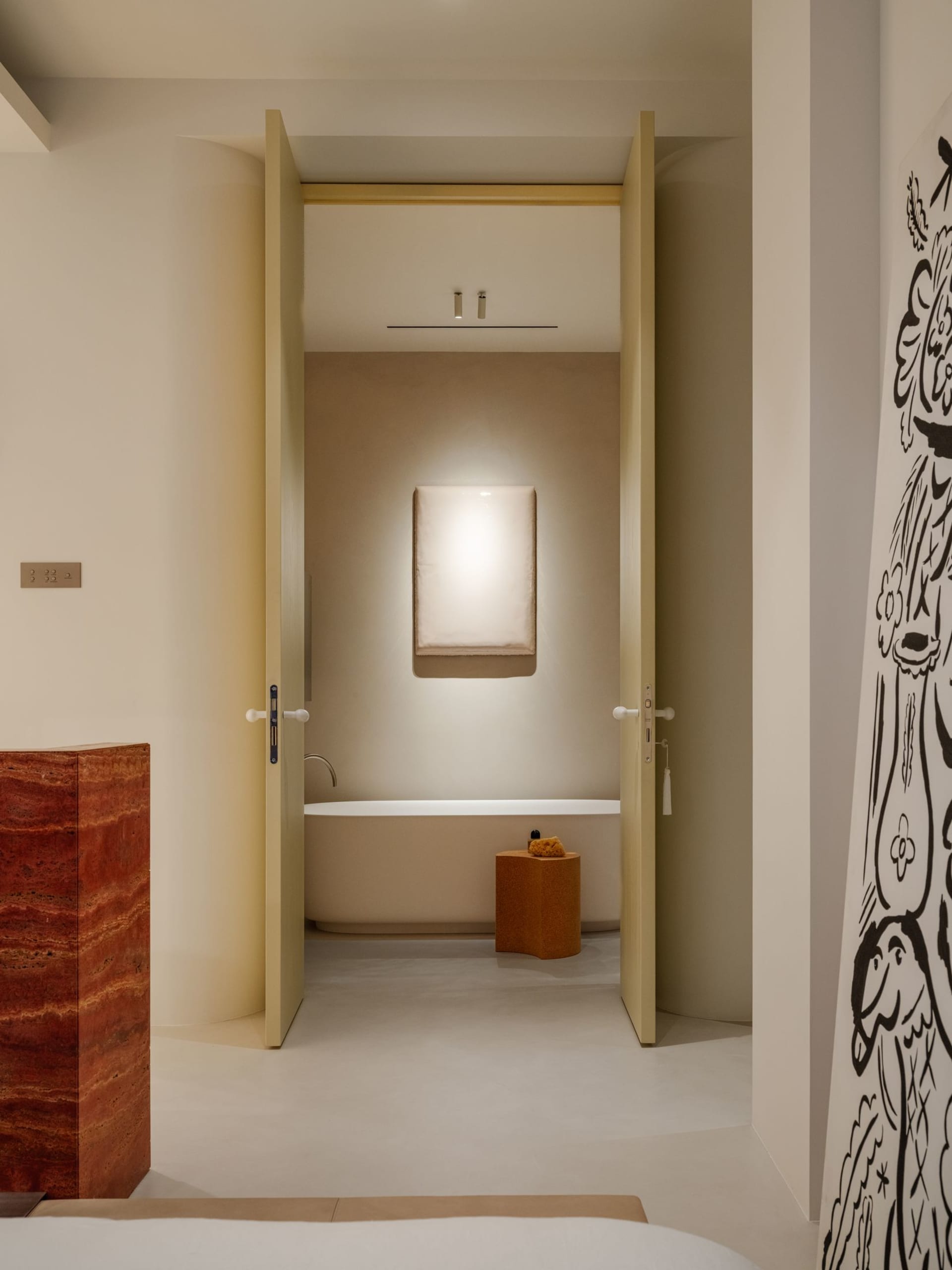 View into a minimalist bathroom with a freestanding white bathtub, a glowing wall sconce, an ochre terrazzo stool, and a red marble block in the foreground.