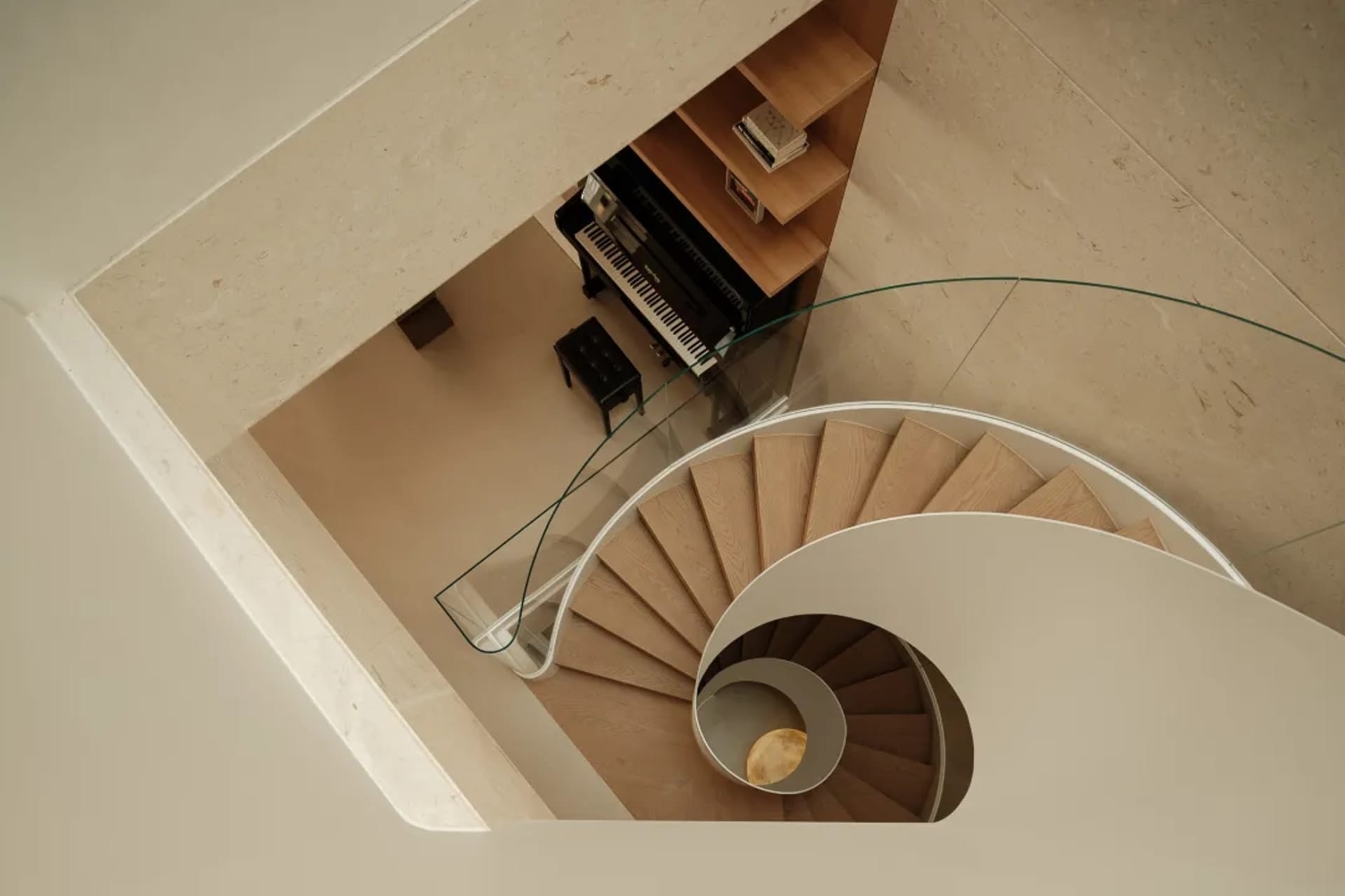 Top-down view of a modern spiral staircase made of light oak with white curved balustrade, glass railing, and a black upright piano nestled in a built-in wooden shelving nook.