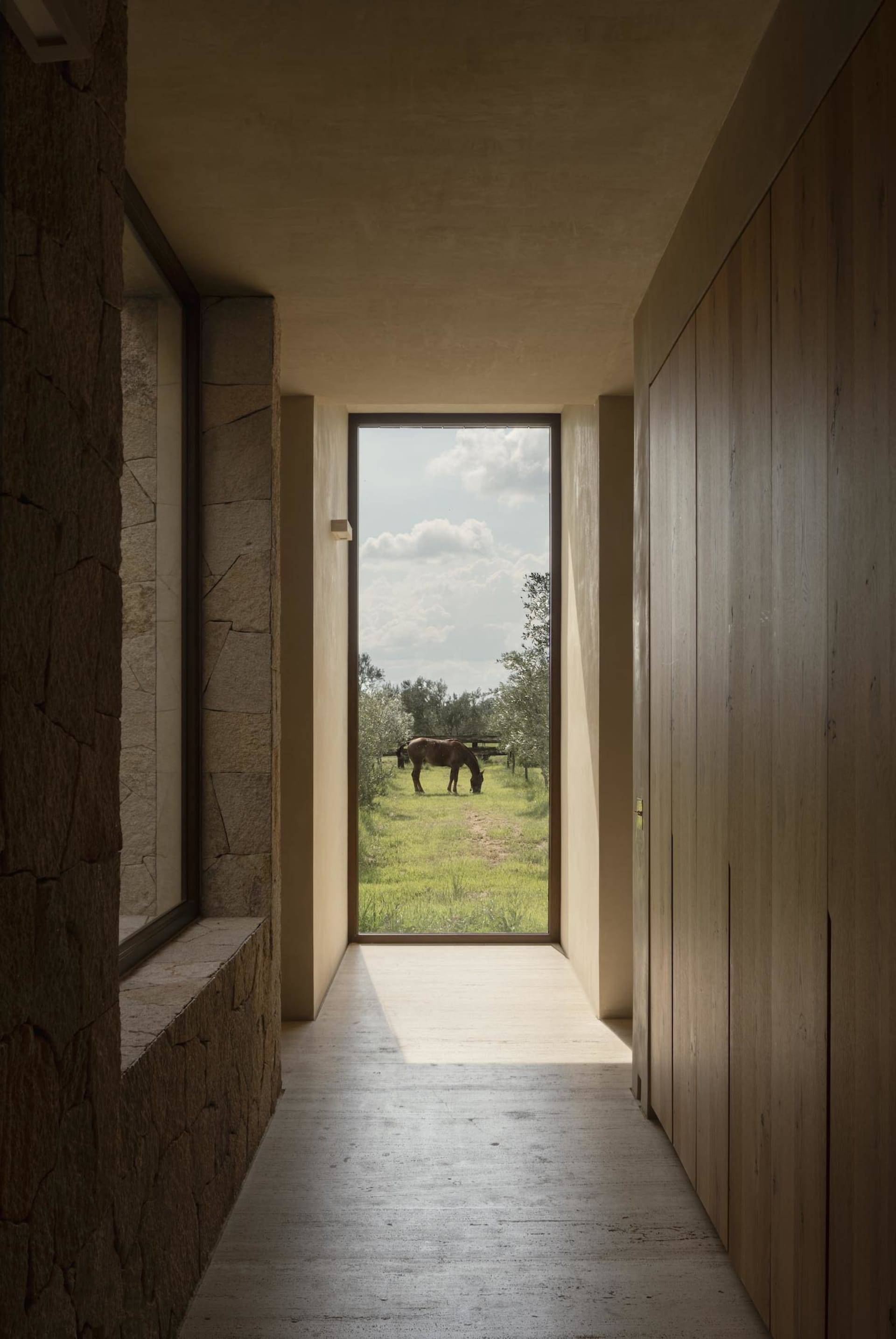 Narrow hallway with rough stone wall on the left, light wood paneling on the right, concrete floor, and a large floor-to-ceiling window at the end revealing a horse grazing in a green pasture under a blue sky.