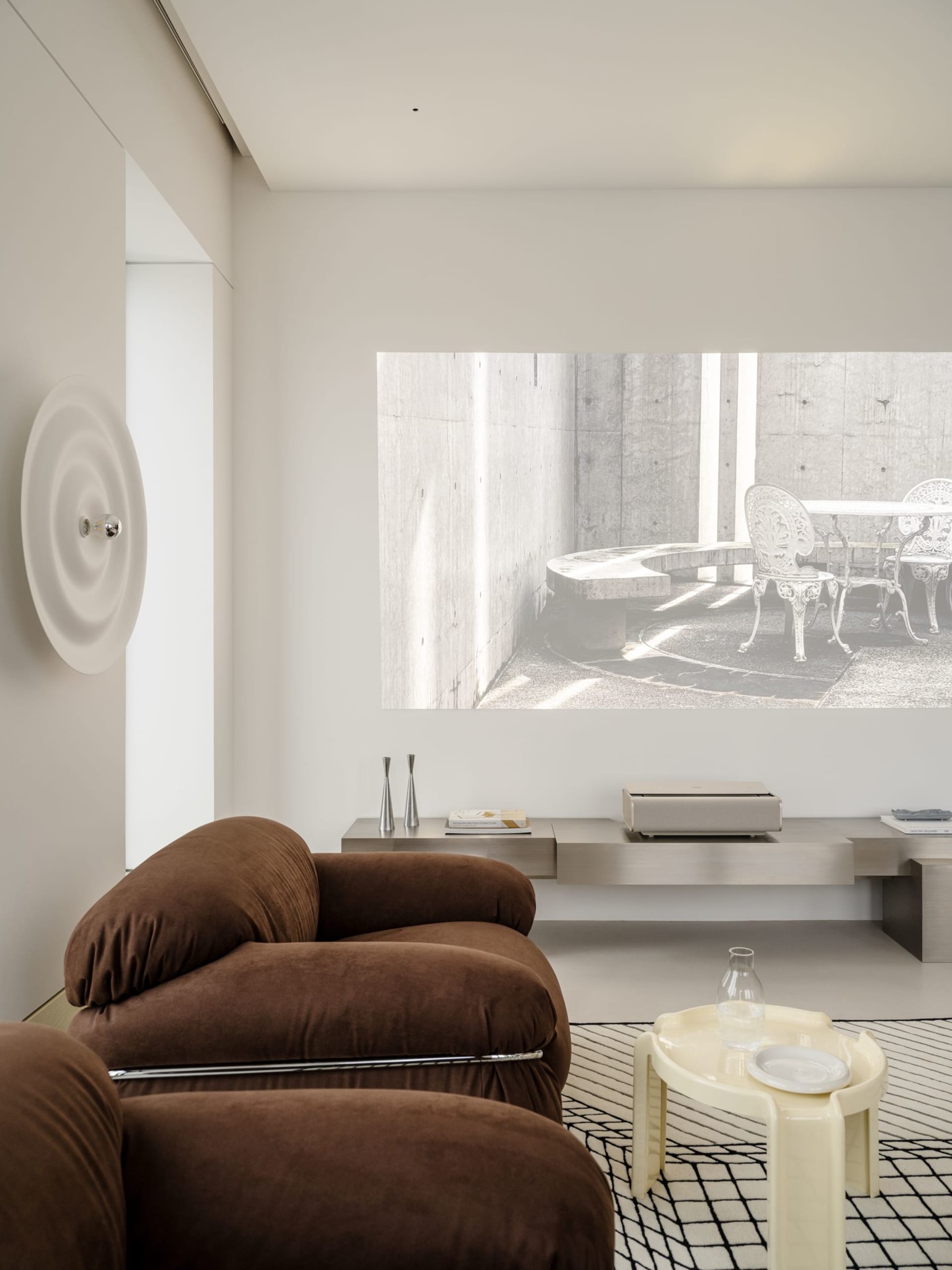 Modern minimalist living room with chocolate brown boucle sofas, cream side table, stainless steel media console, and a large concrete architectural projection on the wall.