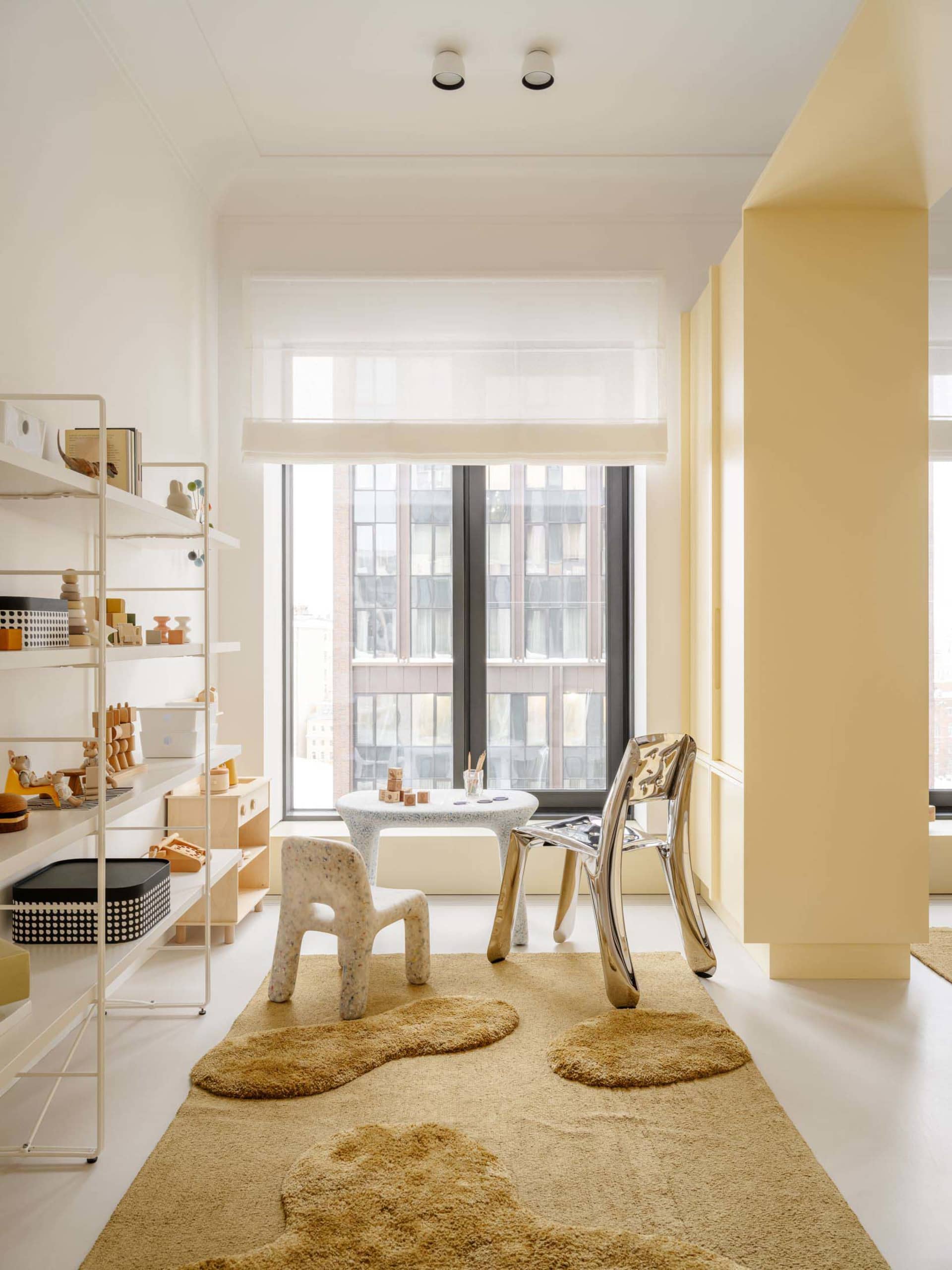Bright modern children's playroom with yellow walls, large window, white shelving, speckled terrazzo table and chair, shiny chrome chair, and irregular beige shaggy rugs