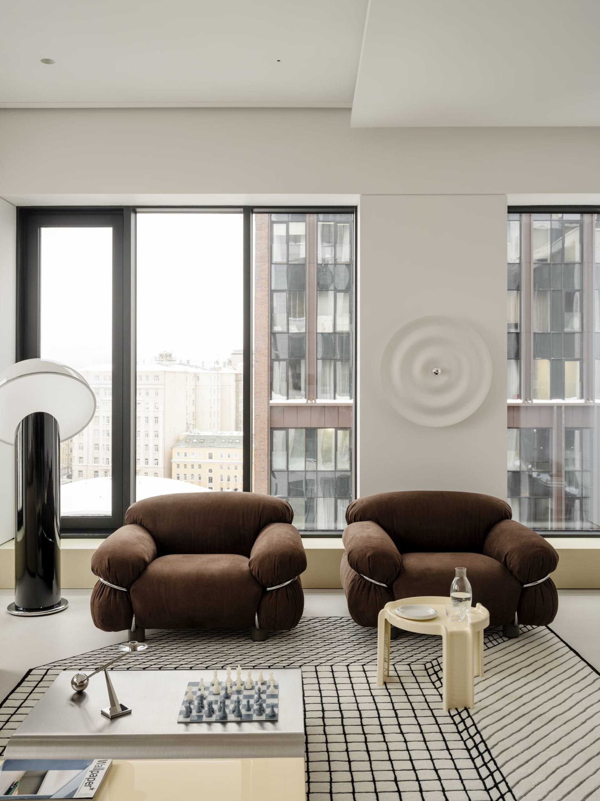 Modern minimalist living room featuring two brown boucle armchairs, a black and white grid-patterned rug, a low white coffee table with a chess set, and large floor-to-ceiling windows overlooking city buildings.