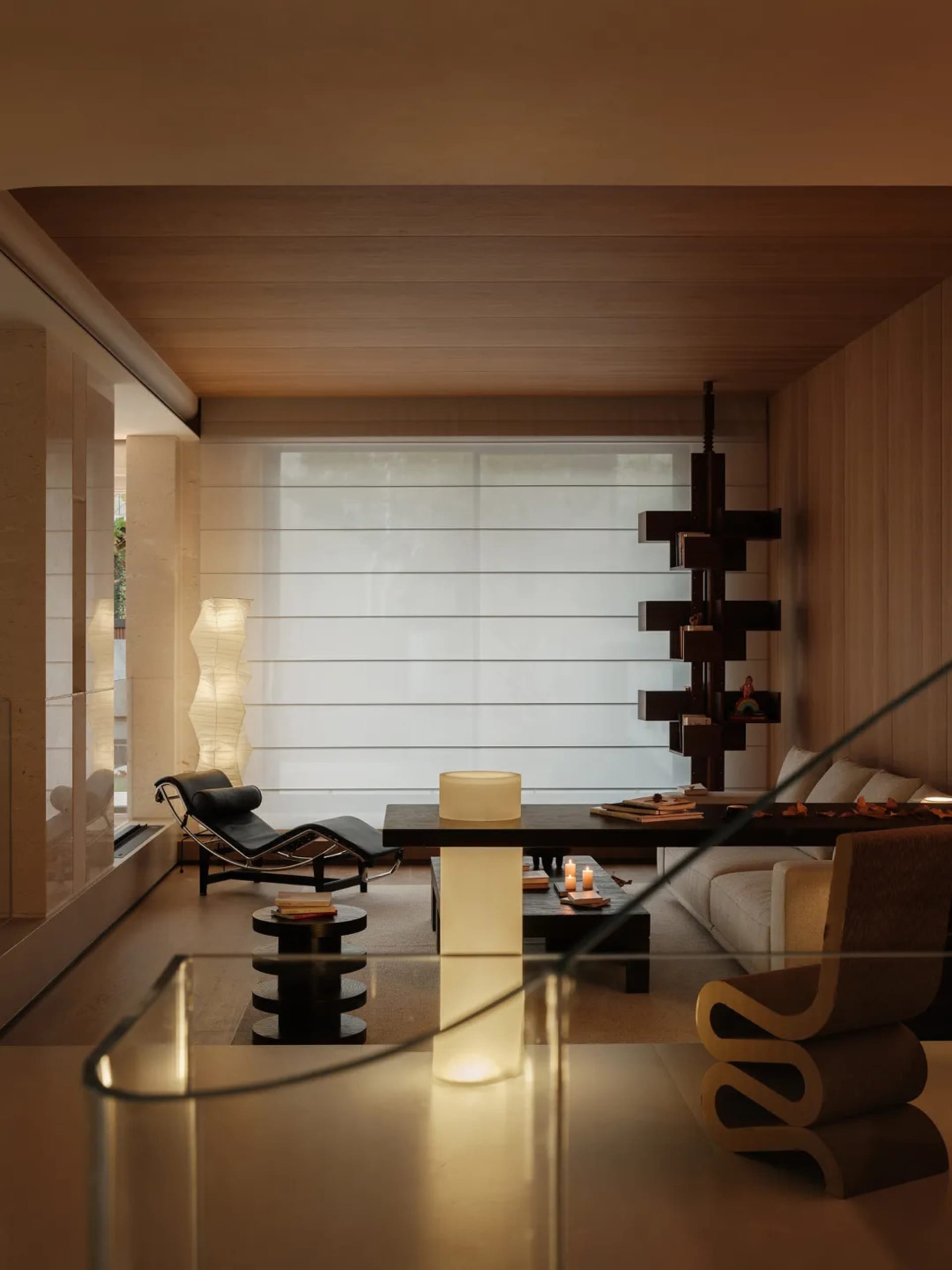 Modern minimalist living room featuring warm wood paneling on walls and ceiling, a black leather chaise lounge, sculptural dark wood bookshelf, layered paper floor lamps, and a low dark coffee table with ambient cylindrical light fixtures.