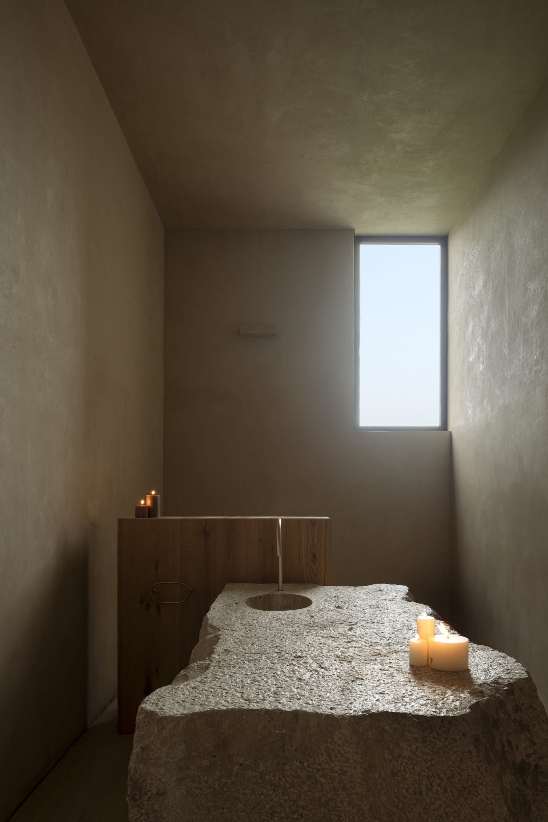 Minimalist bathroom featuring a massive rough-hewn limestone slab with integrated round basin, wooden vanity, brass faucet, and lit candles under a tall vertical window.
