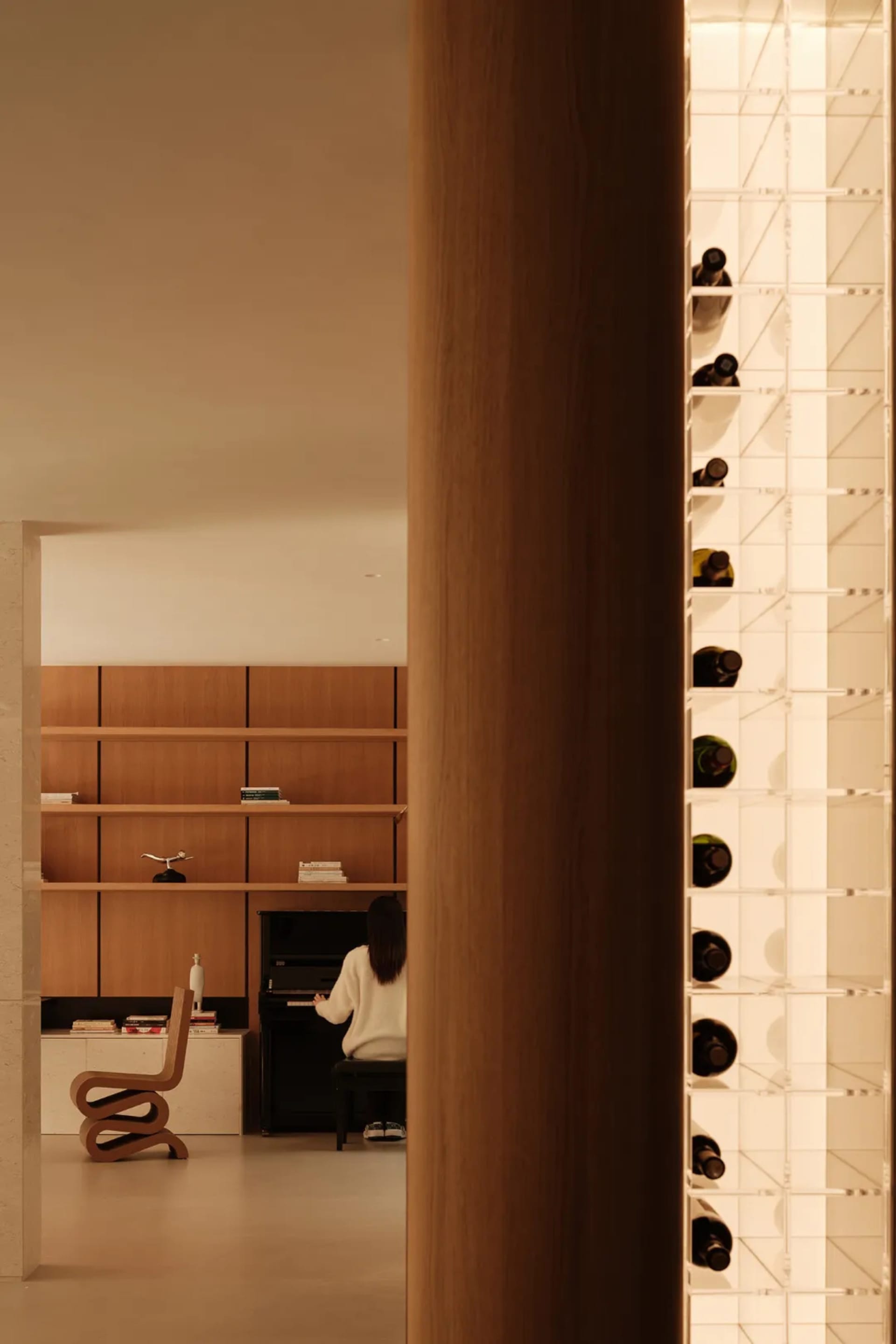 View into a minimalist music room featuring walnut wood shelving, a black upright piano, a sculptural wooden chair, and an illuminated acrylic wine storage wall.