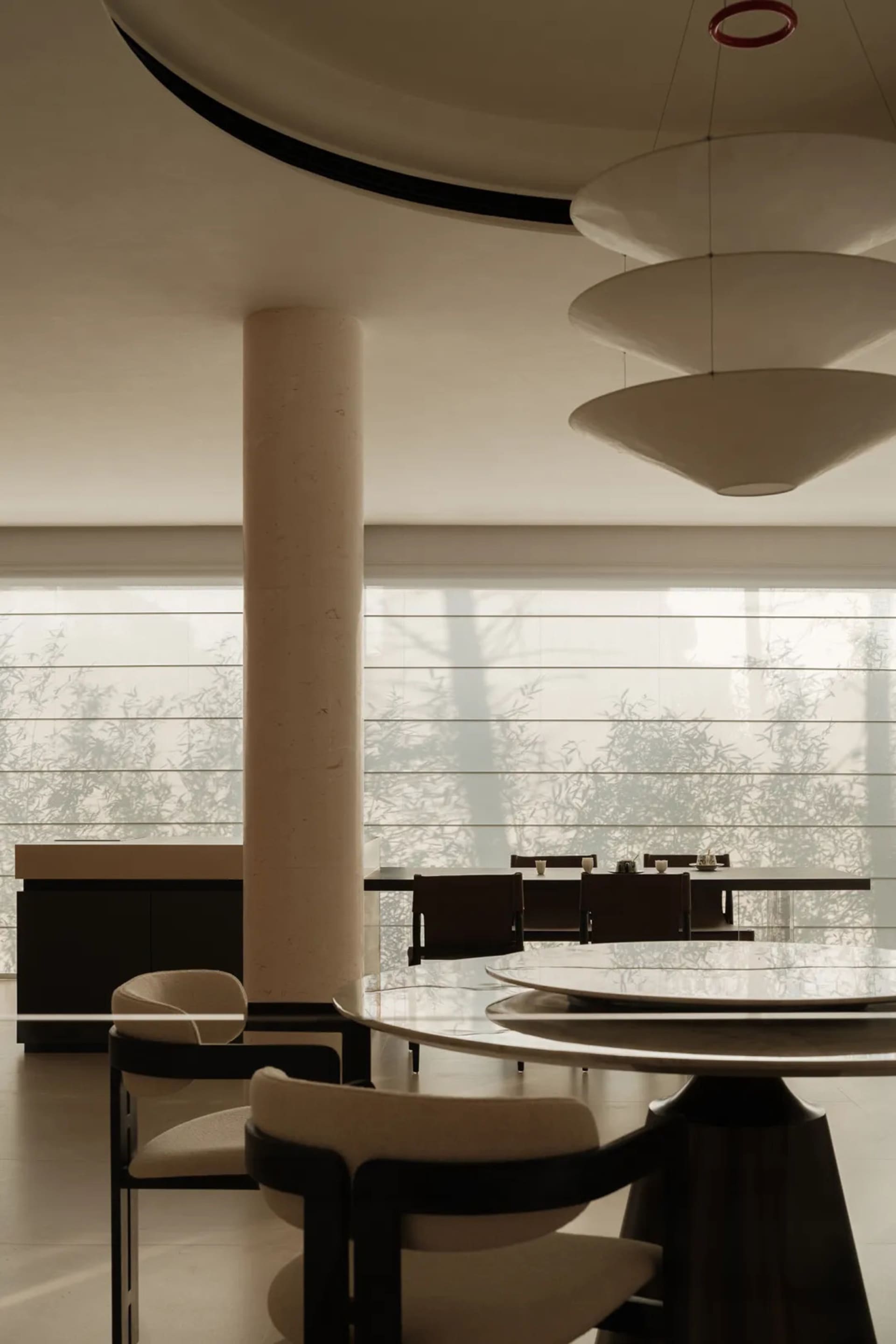 Modern dining room featuring a large round travertine table, curved upholstered chairs, a cylindrical concrete column, and a sculptural three-tiered pendant light with large windows overlooking bamboo in the background.