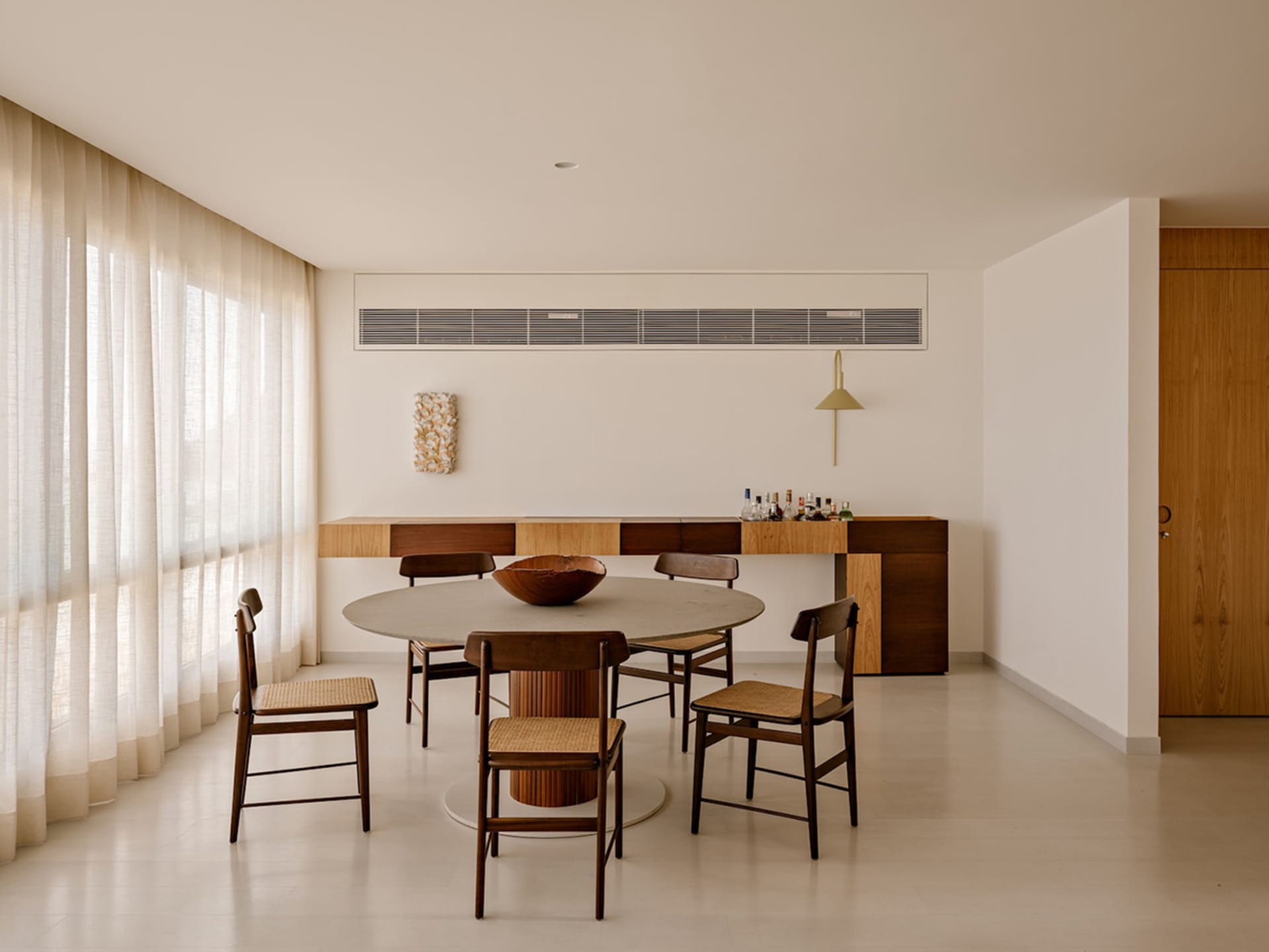 Modern minimalist dining room featuring a large round concrete table surrounded by six dark walnut chairs with woven cane seats, a matching walnut sideboard, and soft sheer curtains allowing diffused daylight.
