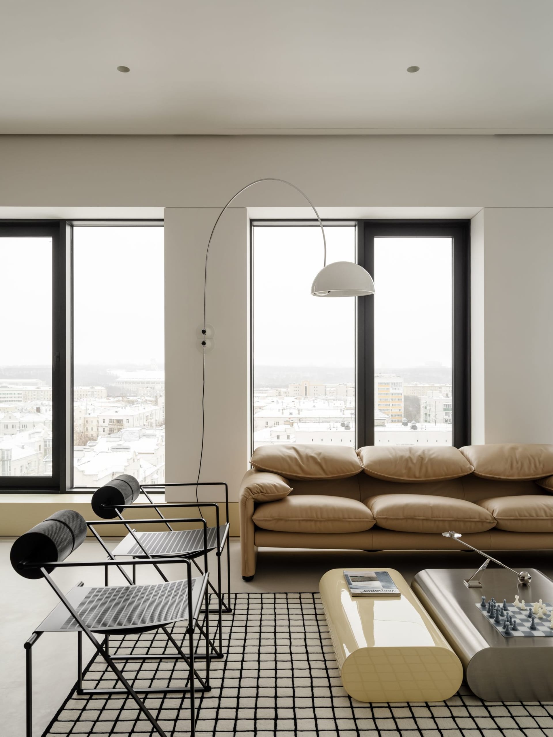 Modern minimalist living room with beige leather sofa, two black wire chairs on a black and white grid rug, glossy curved coffee tables, and large windows showing a snowy city view.