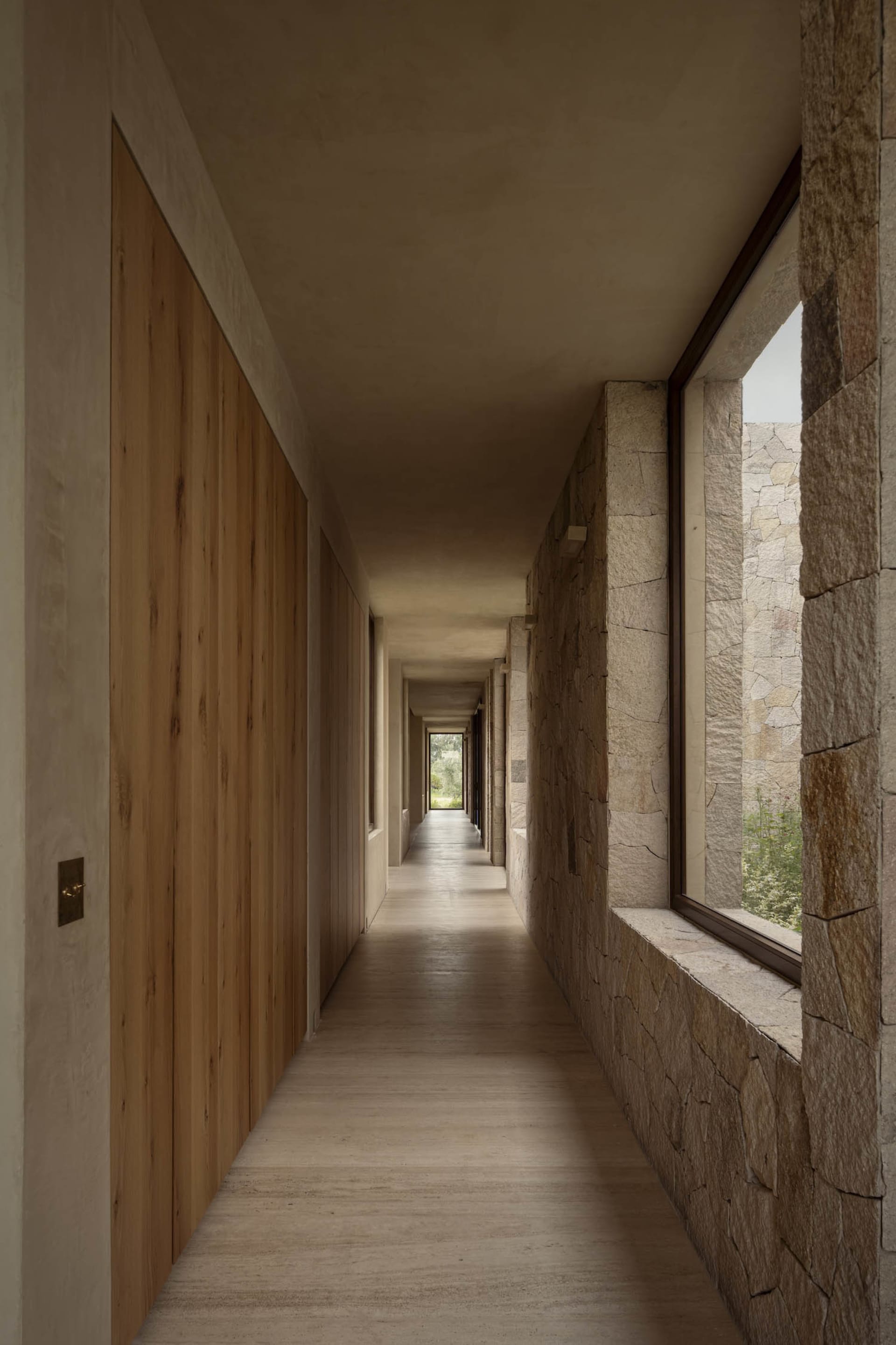 Long minimalist corridor featuring light wood wall paneling on the left and textured natural stone masonry on the right, with large windows allowing daylight to illuminate the concrete floor.