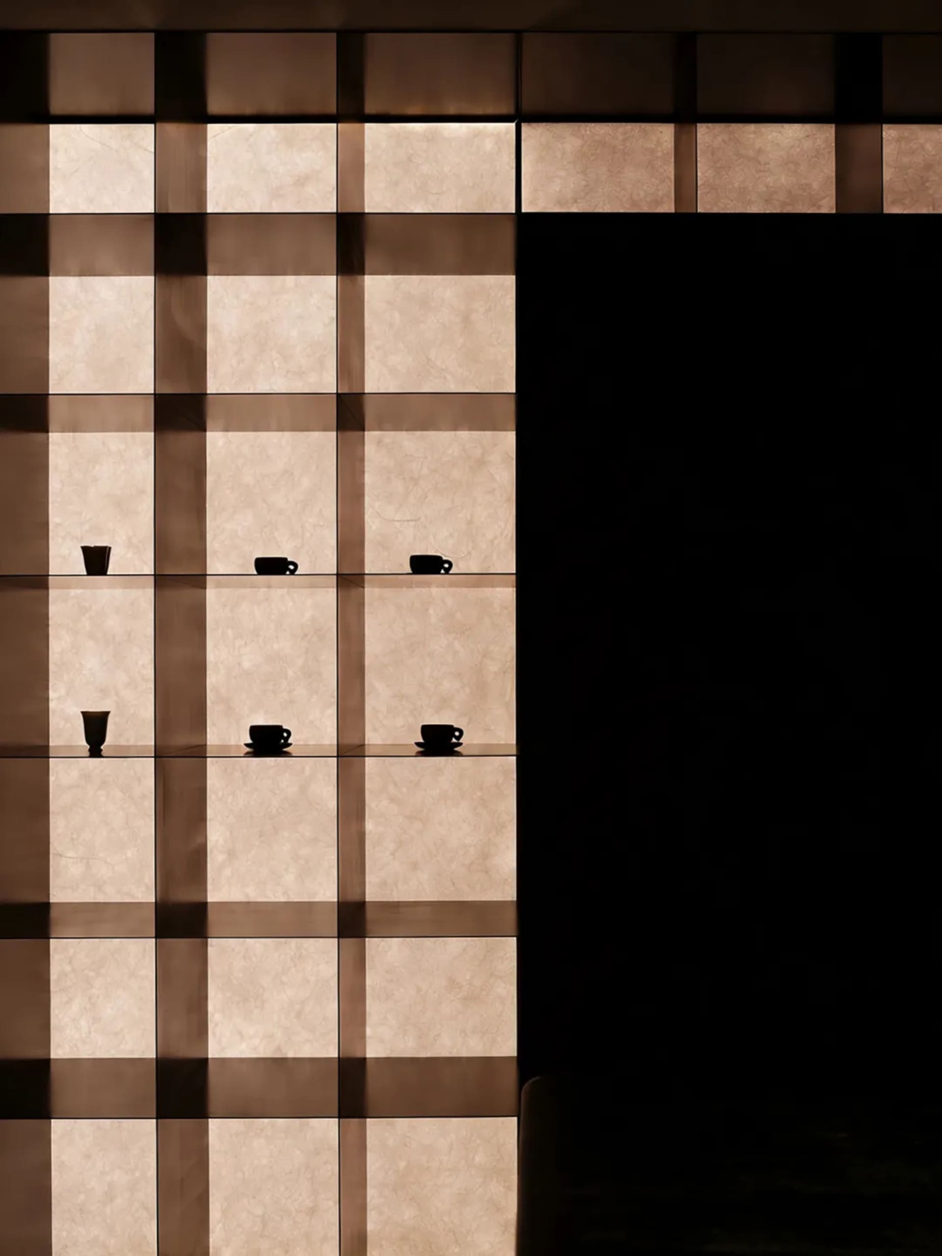 Modern interior wall featuring illuminated honey onyx panels arranged in a grid with floating glass shelves displaying black tableware against a dark background.
