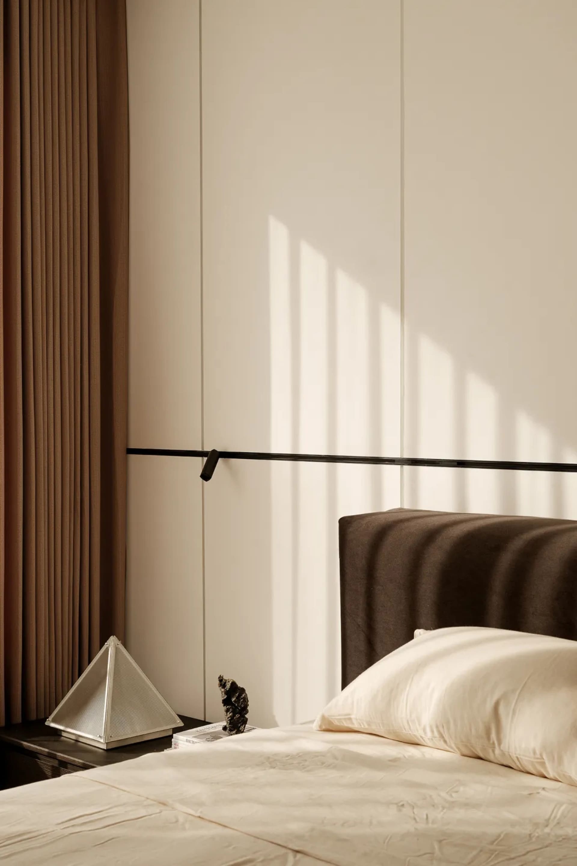 Modern minimalist bedroom corner featuring a dark taupe ribbed fabric headboard, cream bedding, a black floating shelf, and a brown curtain with strong morning sunlight creating striped shadows on the wall.