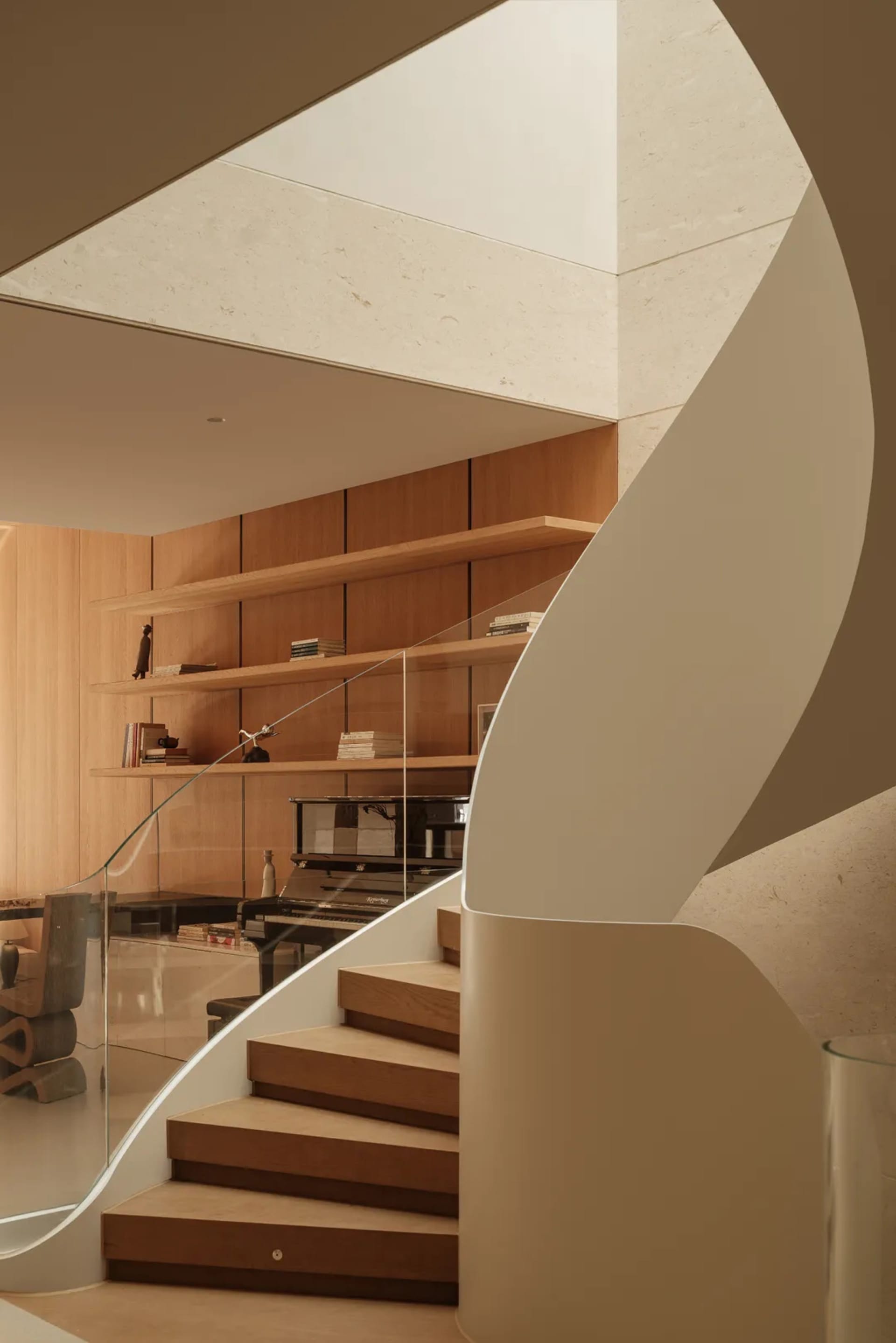Modern curved oak staircase with white curved wall detail, floating wooden shelves, glass railing, and a grand piano visible in the background of a minimalist living room.
