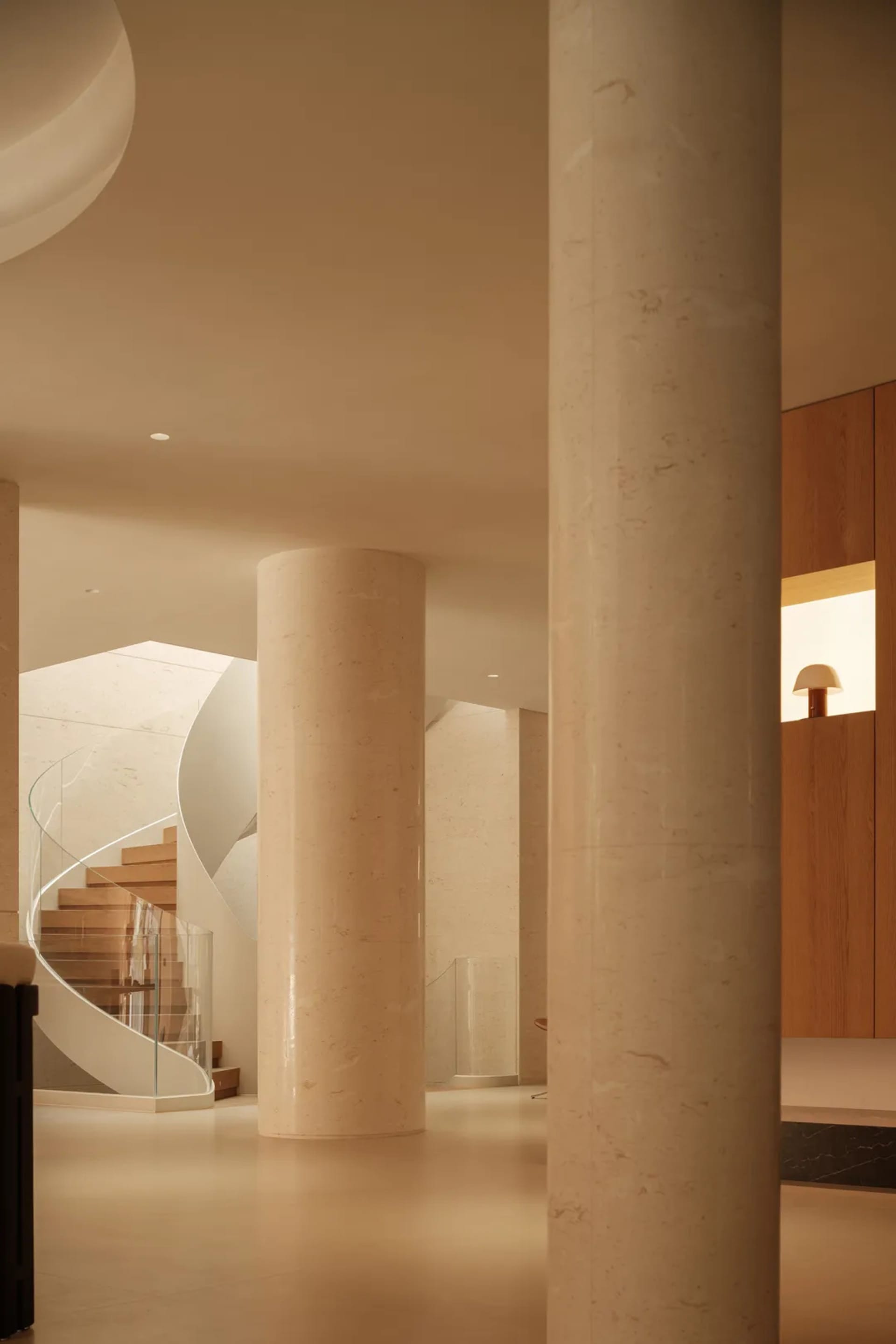 Modern interior featuring large cylindrical travertine columns, a sculptural curved wooden staircase with glass railing, and warm ambient lighting.