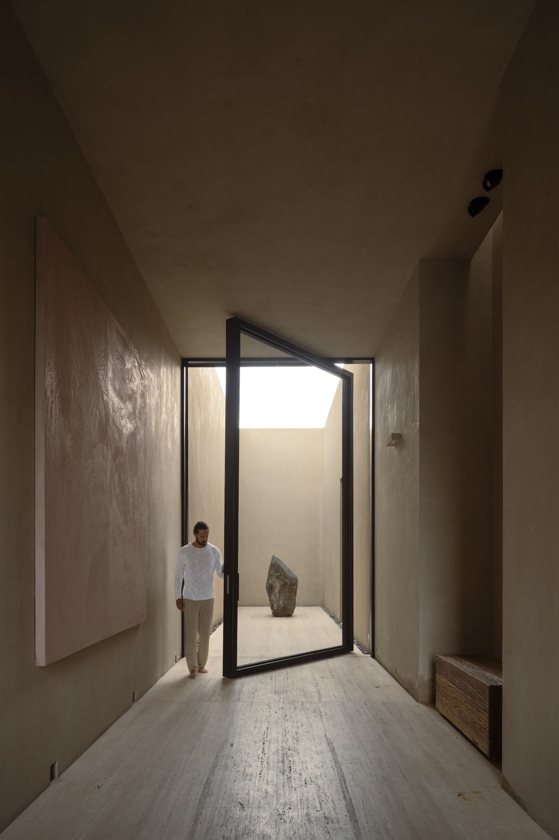 Minimalist concrete hallway with large abstract wall art, black-framed pivot door opening to a sunlit courtyard containing a large natural stone sculpture.