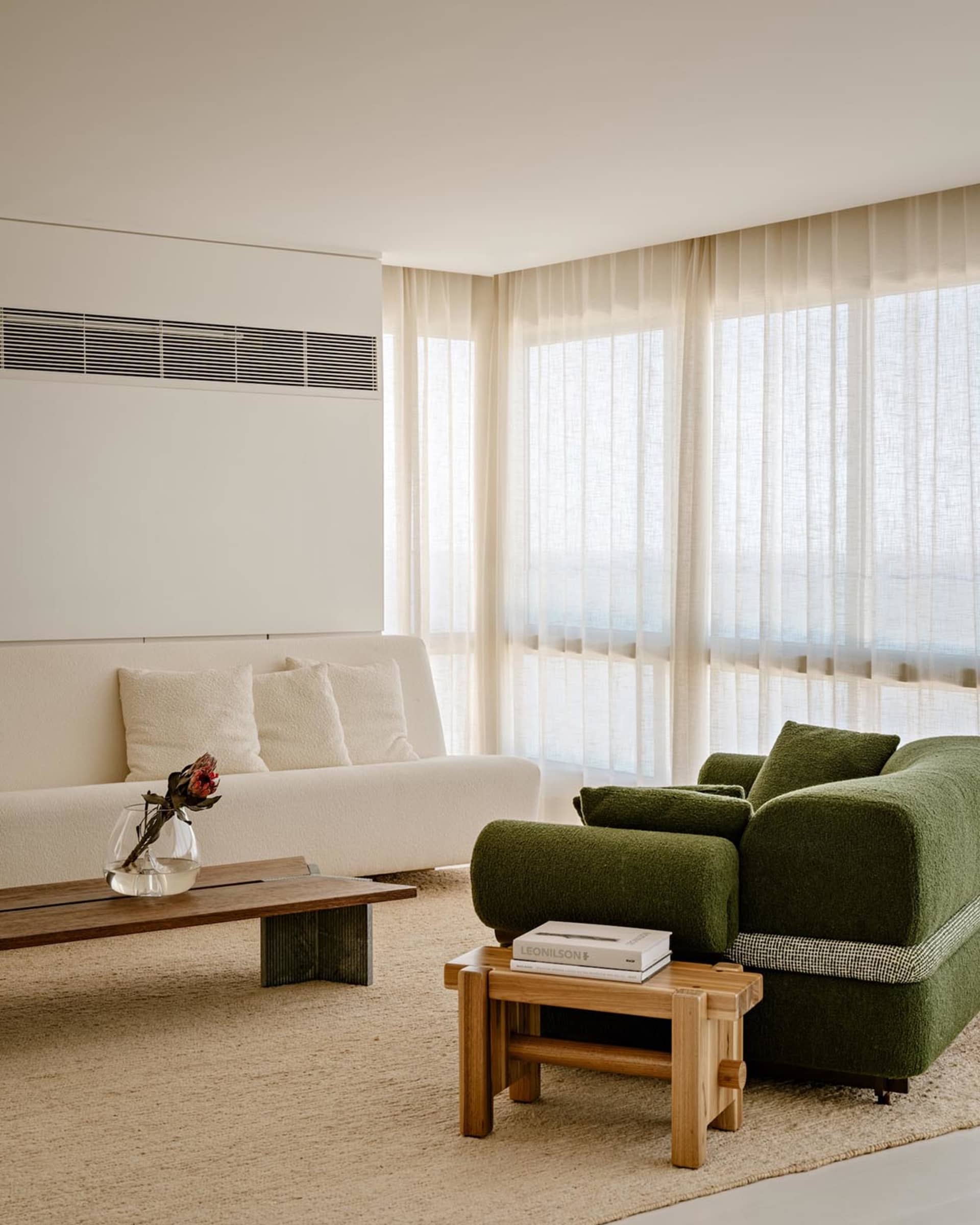Modern minimalist living room featuring a moss green boucle sofa, off-white bouclé sofa, low oak coffee tables, and large windows with sheer curtains.