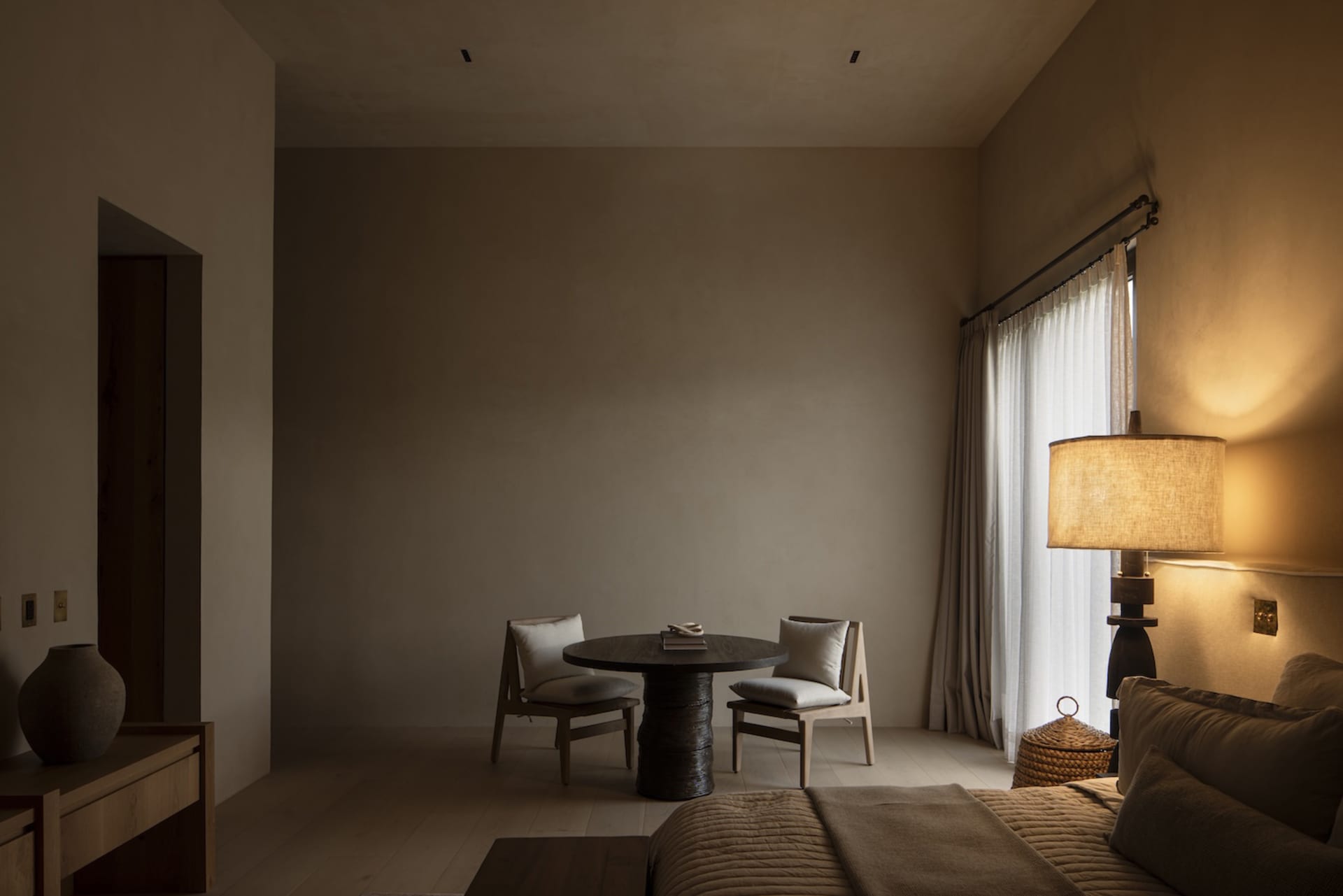 Modern minimalist bedroom interior featuring a dark oak round table with two upholstered chairs, a tall woven floor lamp casting warm light, neutral beige walls, and a bed with textured linen bedding.