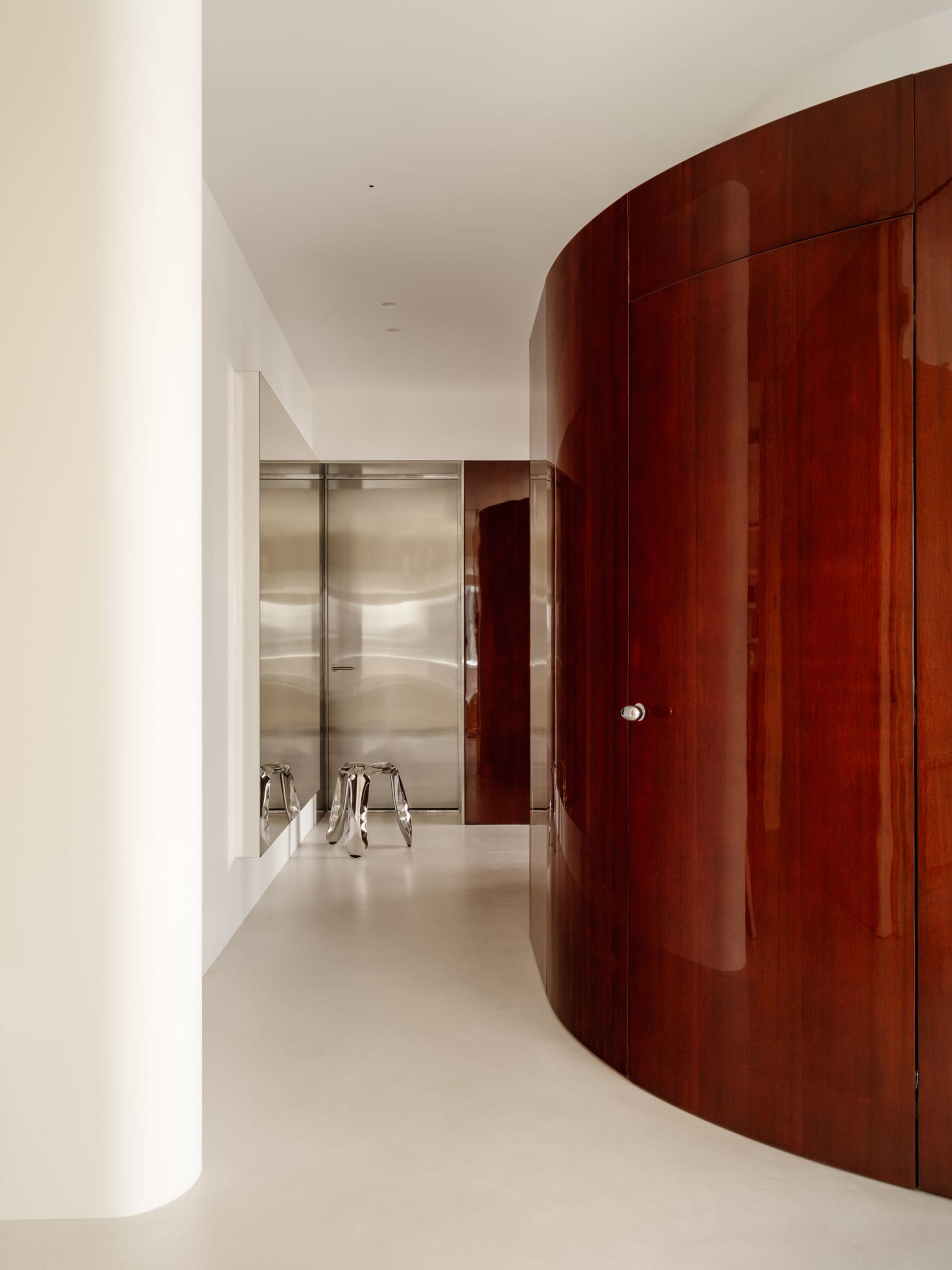 Modern minimalist hallway featuring a large curved wardrobe in rich rosewood finish, white walls and floor, with stainless steel doors and sculptural metallic stools in the background.
