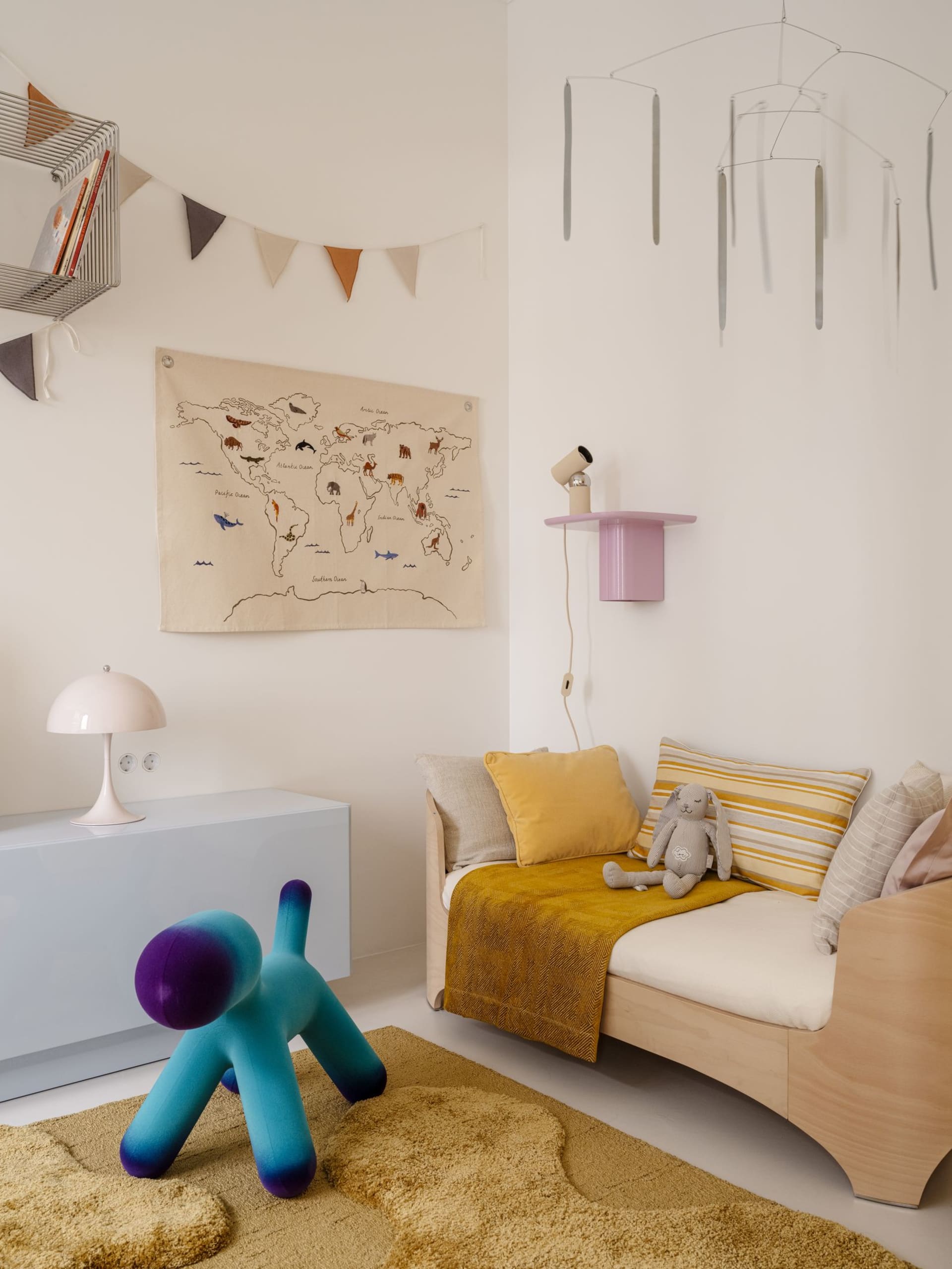 Bright children's room corner with a wooden daybed, mustard yellow textiles, a colorful gradient dog-shaped stool, world map wall hanging, and modern pendant light.