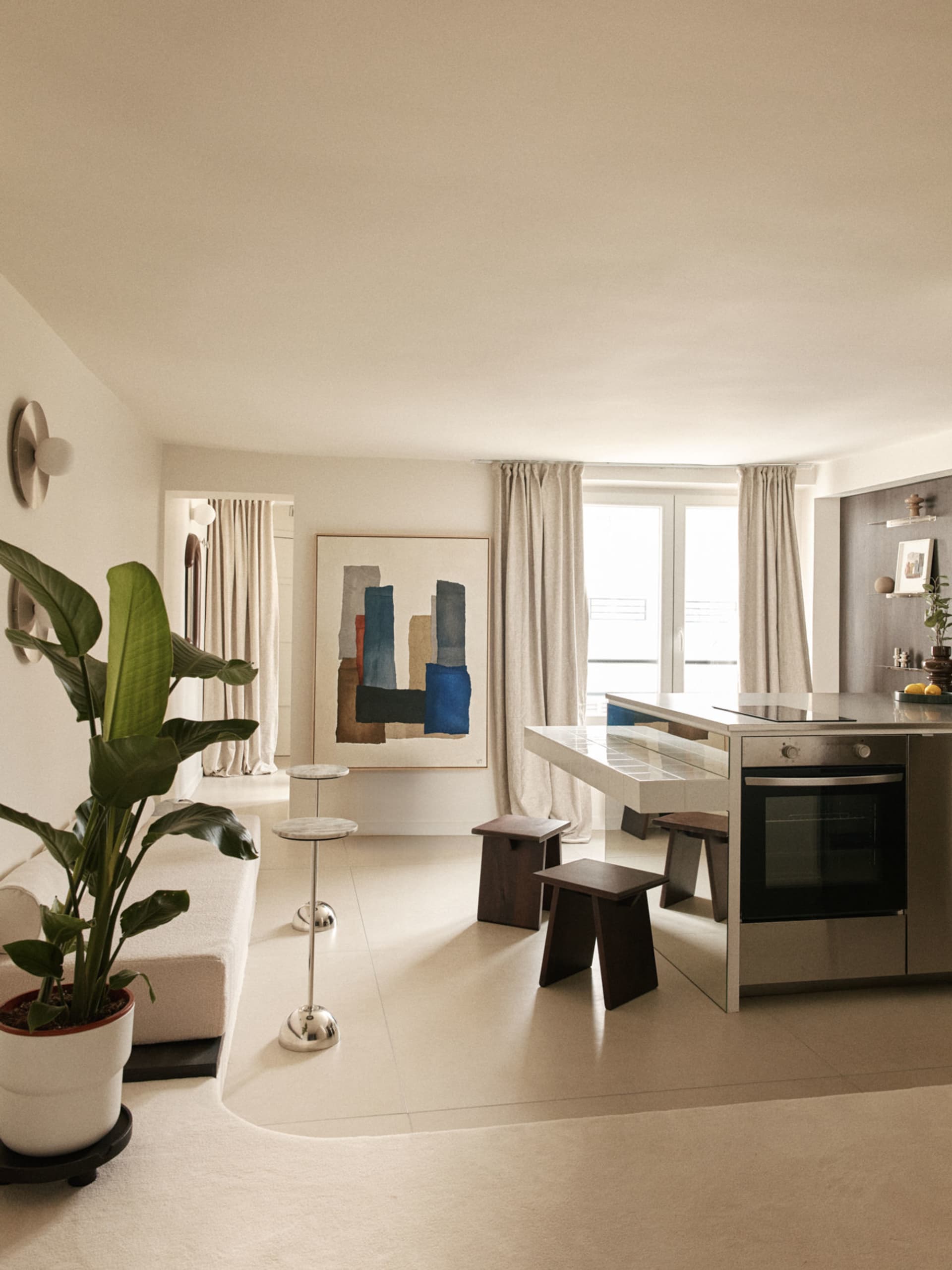 Modern open-plan living and kitchen area with large abstract painting, white marble kitchen island, dark wood stools, cream sofa, and large potted plant.