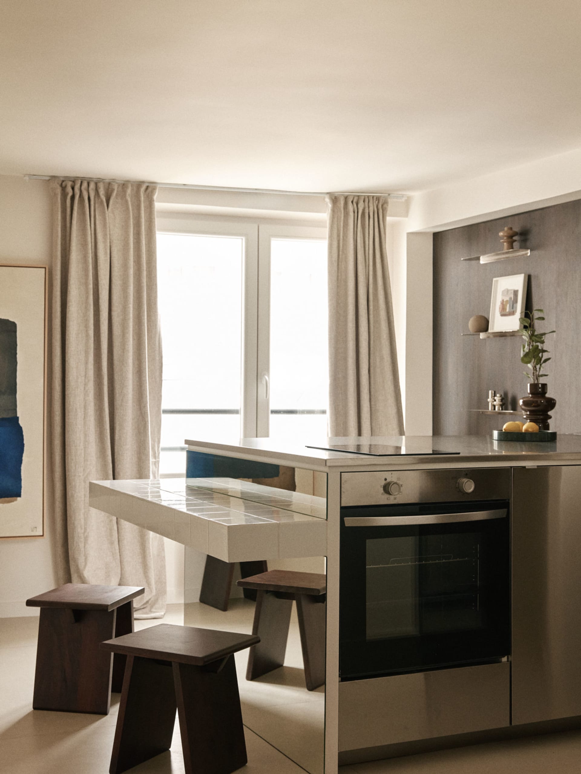 Modern minimalist kitchen featuring a white tiled breakfast bar, built-in oven, walnut wood stools, and large windows with linen curtains allowing abundant natural light.