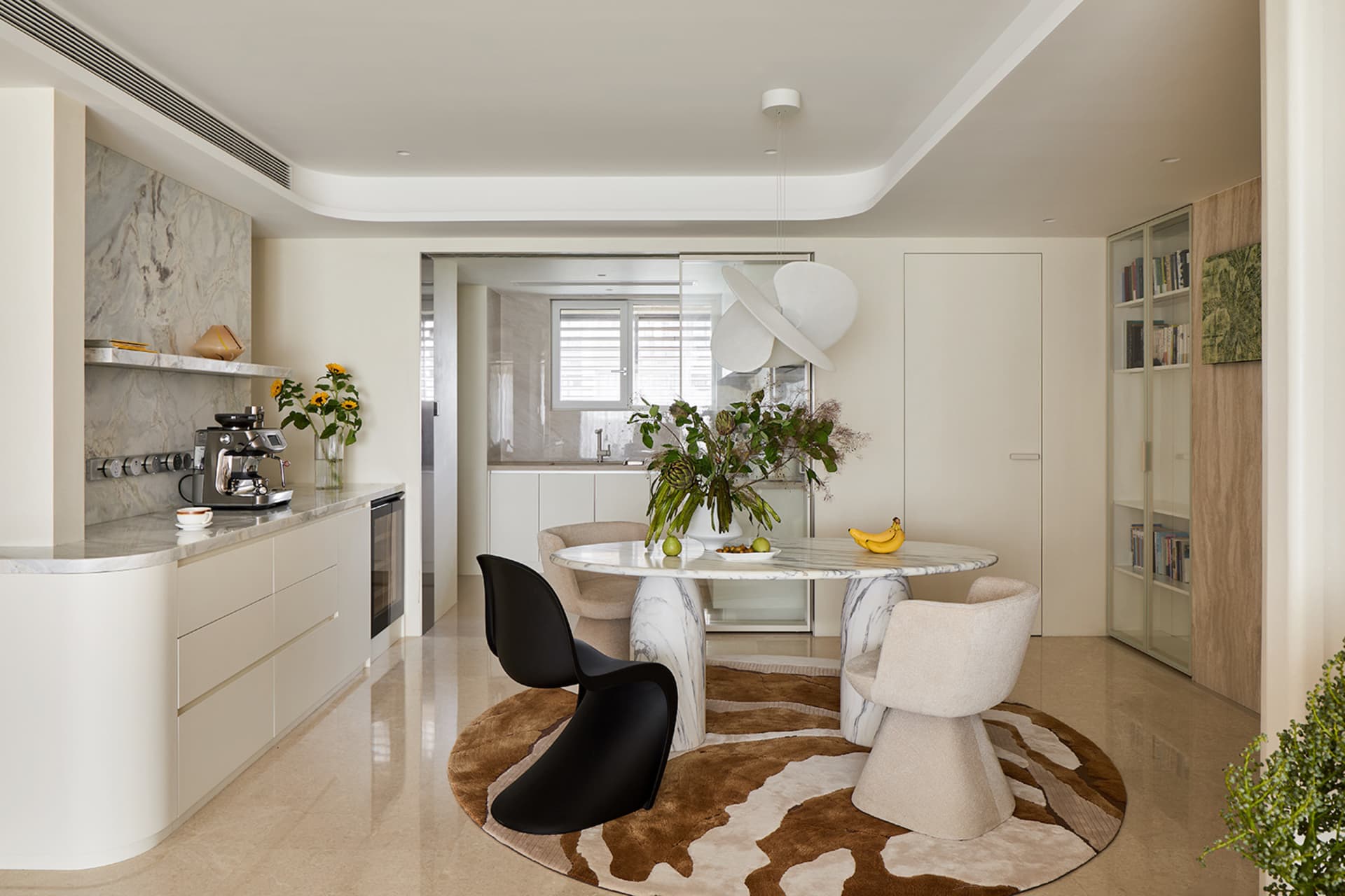 Modern dining area featuring a round Calacatta marble table, black Panton chair, cream boucle chairs, and a brown-and-white cowhide rug in a bright open-plan kitchen and living space.