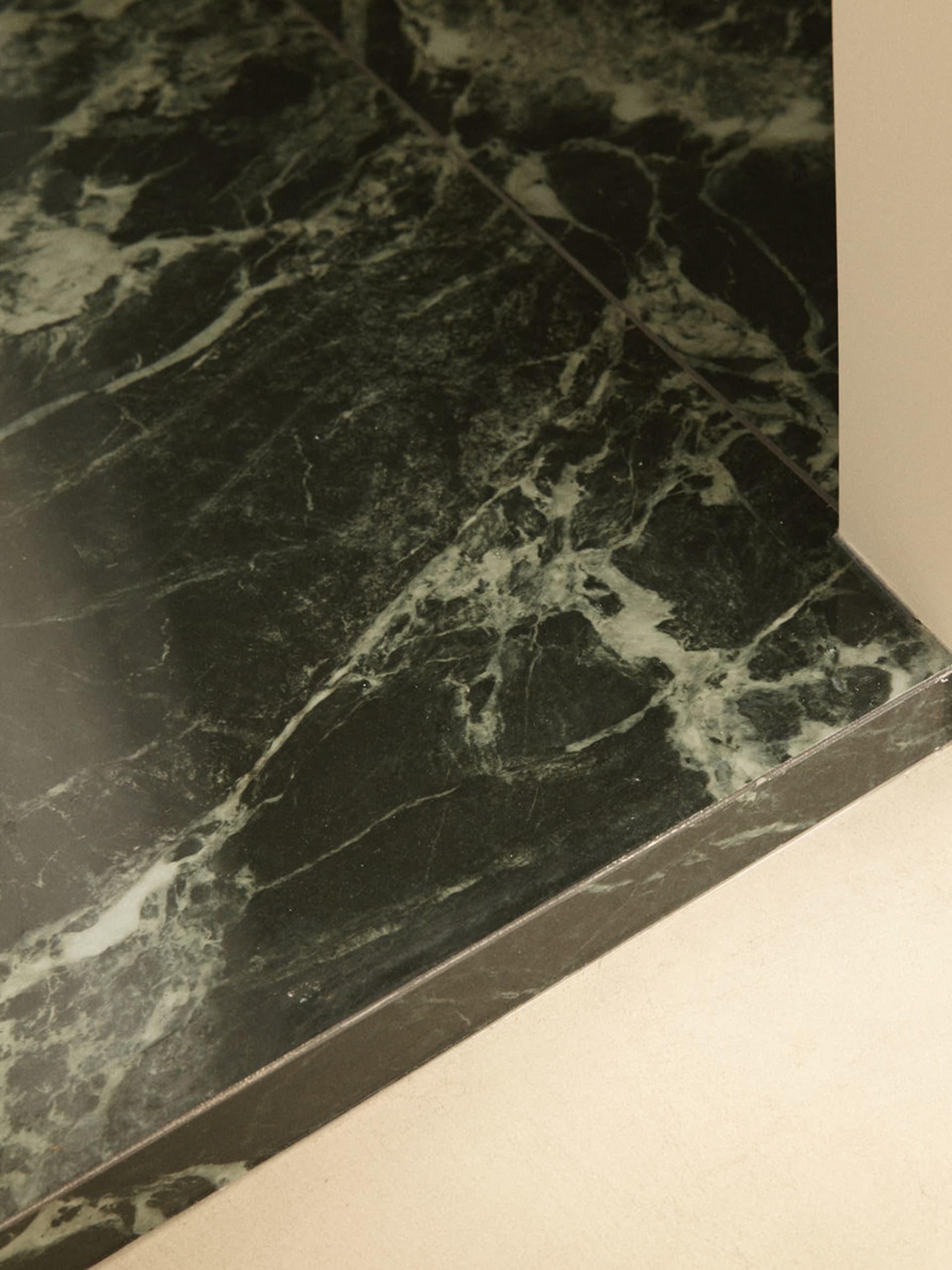 Close-up photograph of dark green marble flooring with prominent white and grey veining, showing the polished surface and edge detail against a light wall and baseboard.