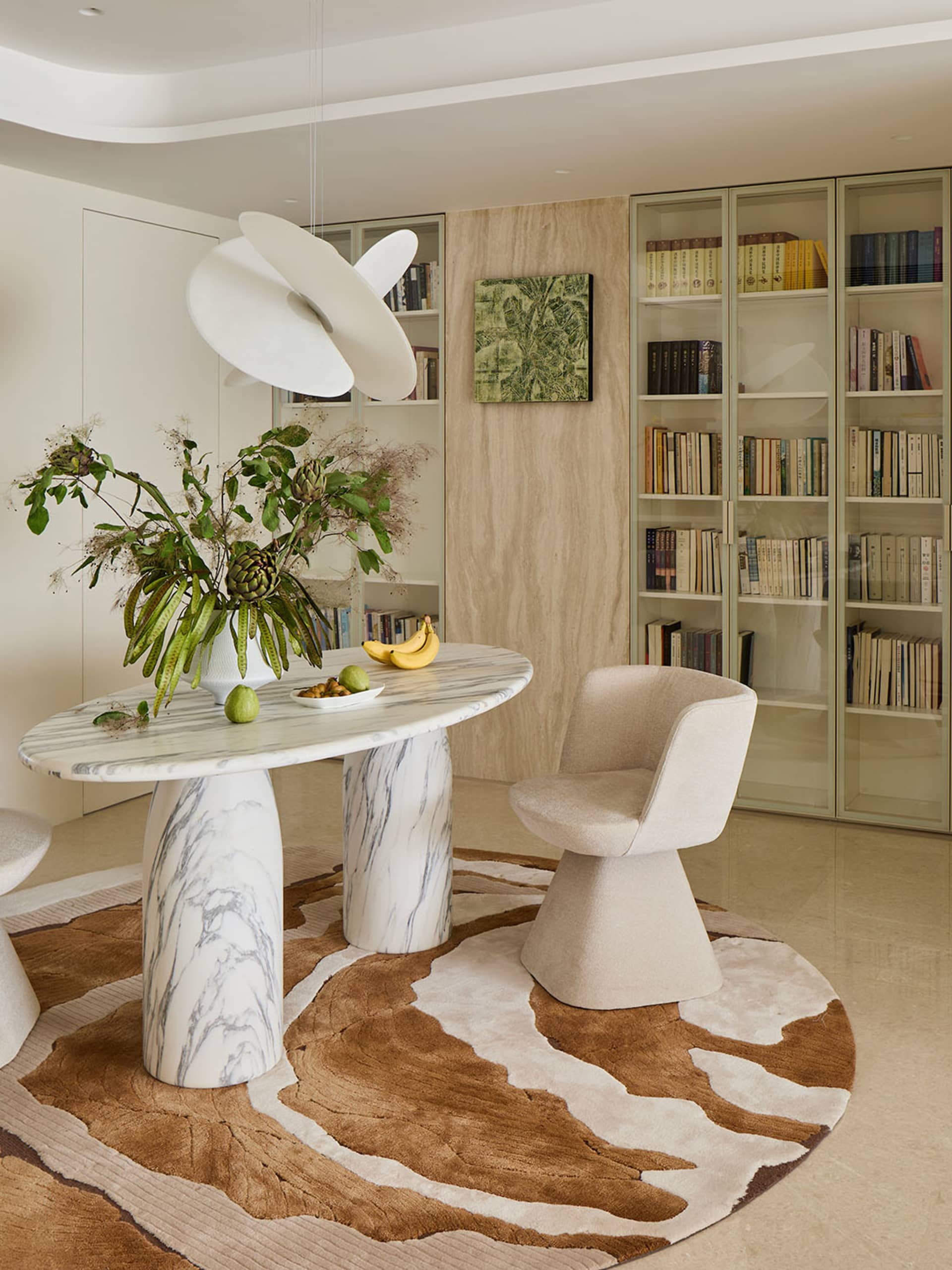 Modern dining area featuring a round Calacatta marble table on sculptural bases, a cream bouclé swivel chair, a large white pendant lamp, and floor-to-ceiling built-in bookshelves with light wood paneling.