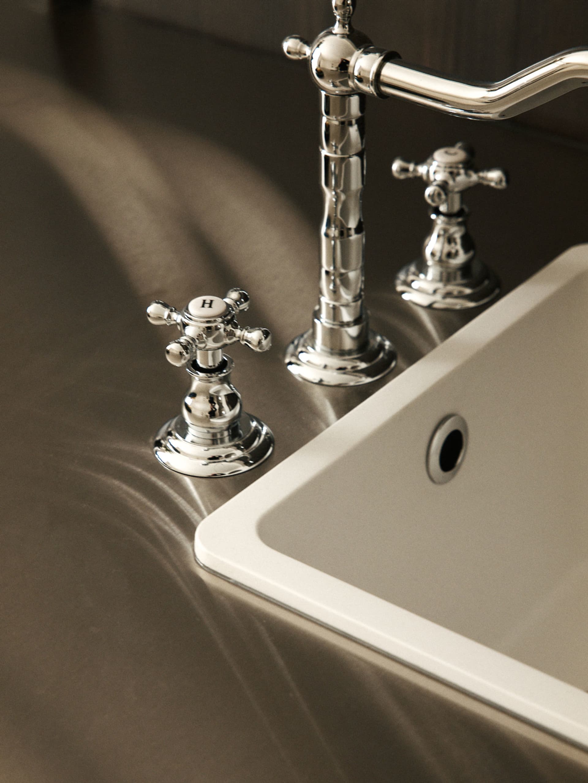 Close-up of a polished chrome bathroom faucet with white ceramic cross handles marked with H and C, mounted on a smooth stone countertop next to a white ceramic sink.