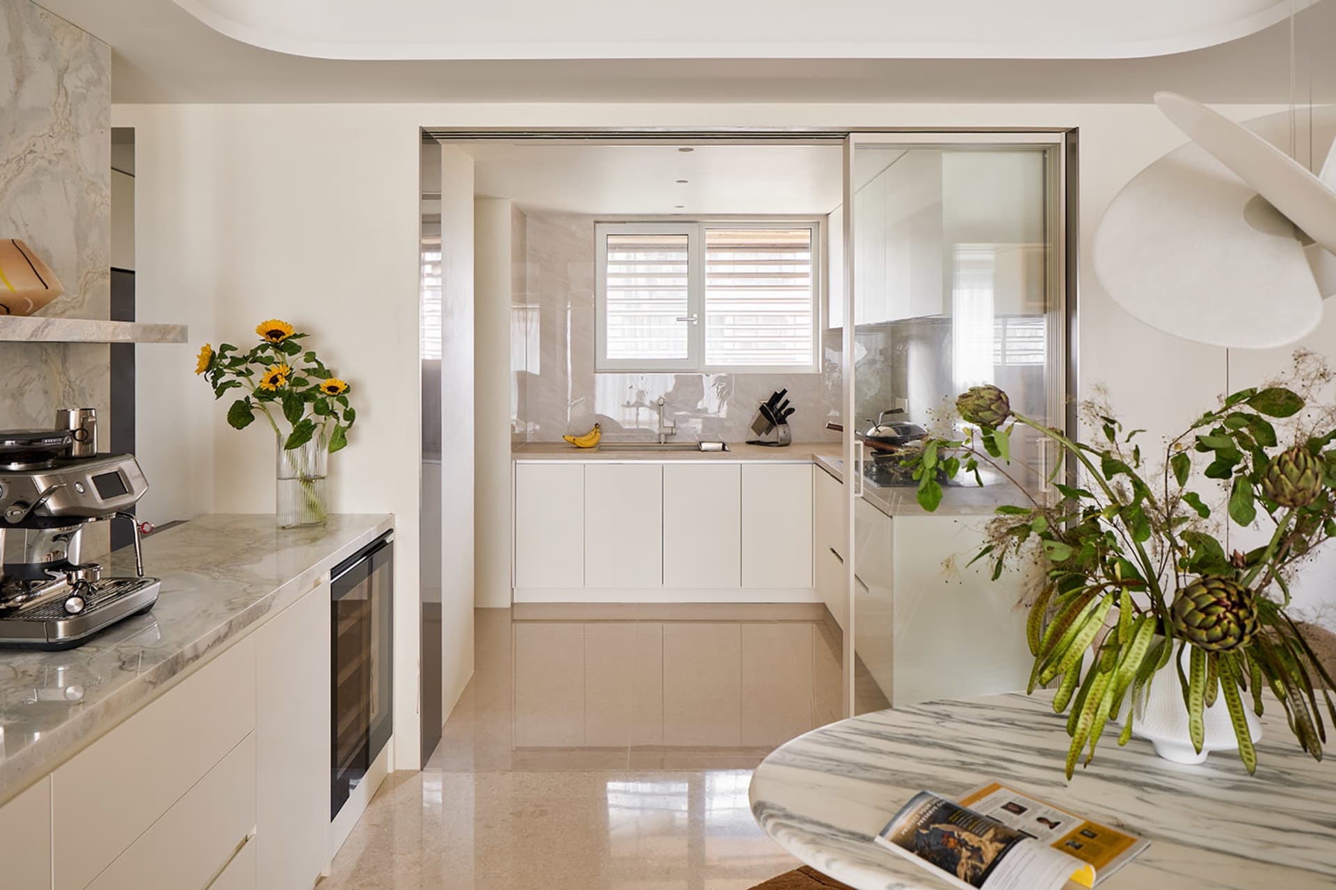 Modern open-concept kitchen with white cabinetry, Calacatta marble countertops and island, stainless steel espresso machine, and large white pendant light.