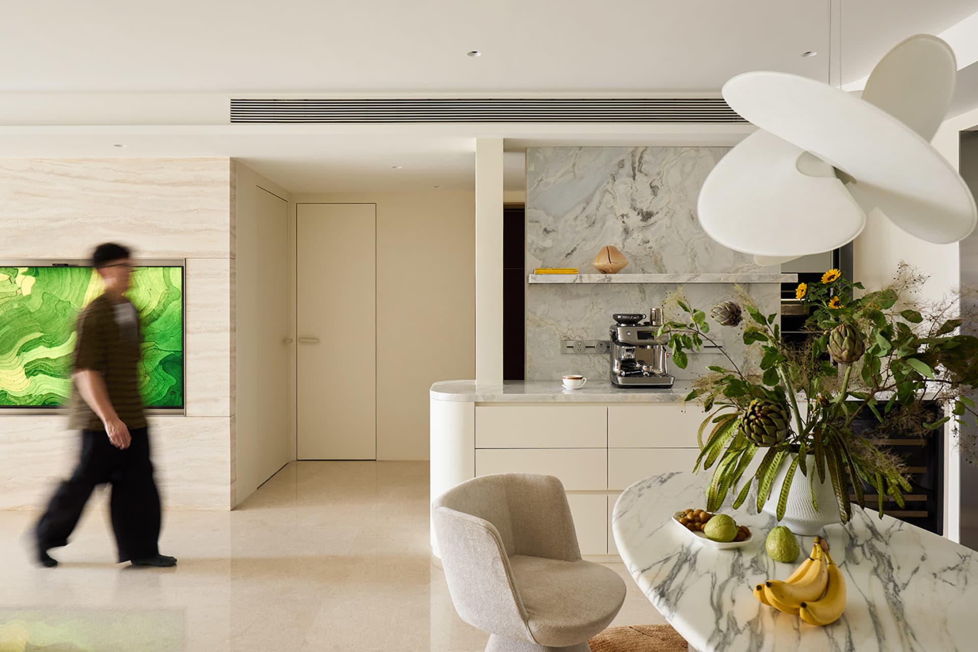 Modern open-plan kitchen and living area featuring a curved Calacatta marble island, sculptural white pendant light, built-in coffee station against a marble backsplash, and a beige boucle swivel chair.