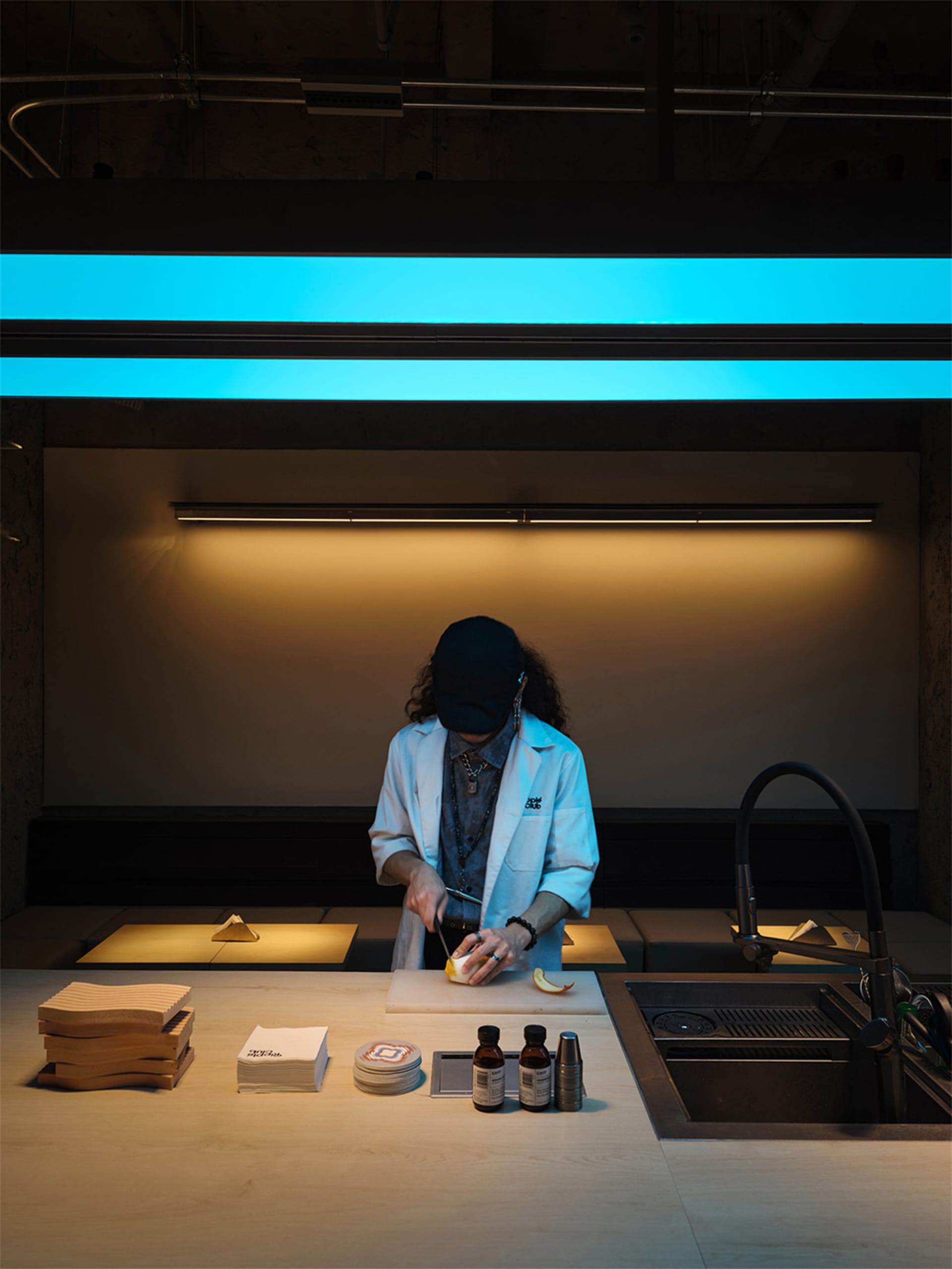A person with curly hair wearing a white chef coat and black cap stands at a concrete kitchen counter, slicing a lemon under cool cyan and warm ambient lighting in an industrial-style commercial kitchen.