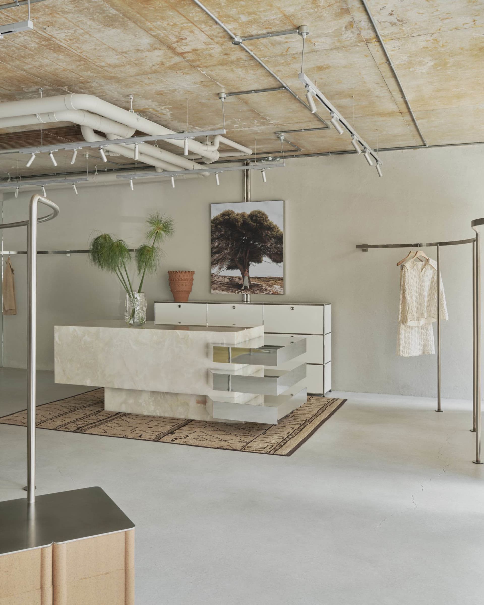 Modern minimalist boutique interior featuring a large travertine stone central island, white modular cabinetry, industrial track lighting, and clothing racks under an exposed concrete ceiling.