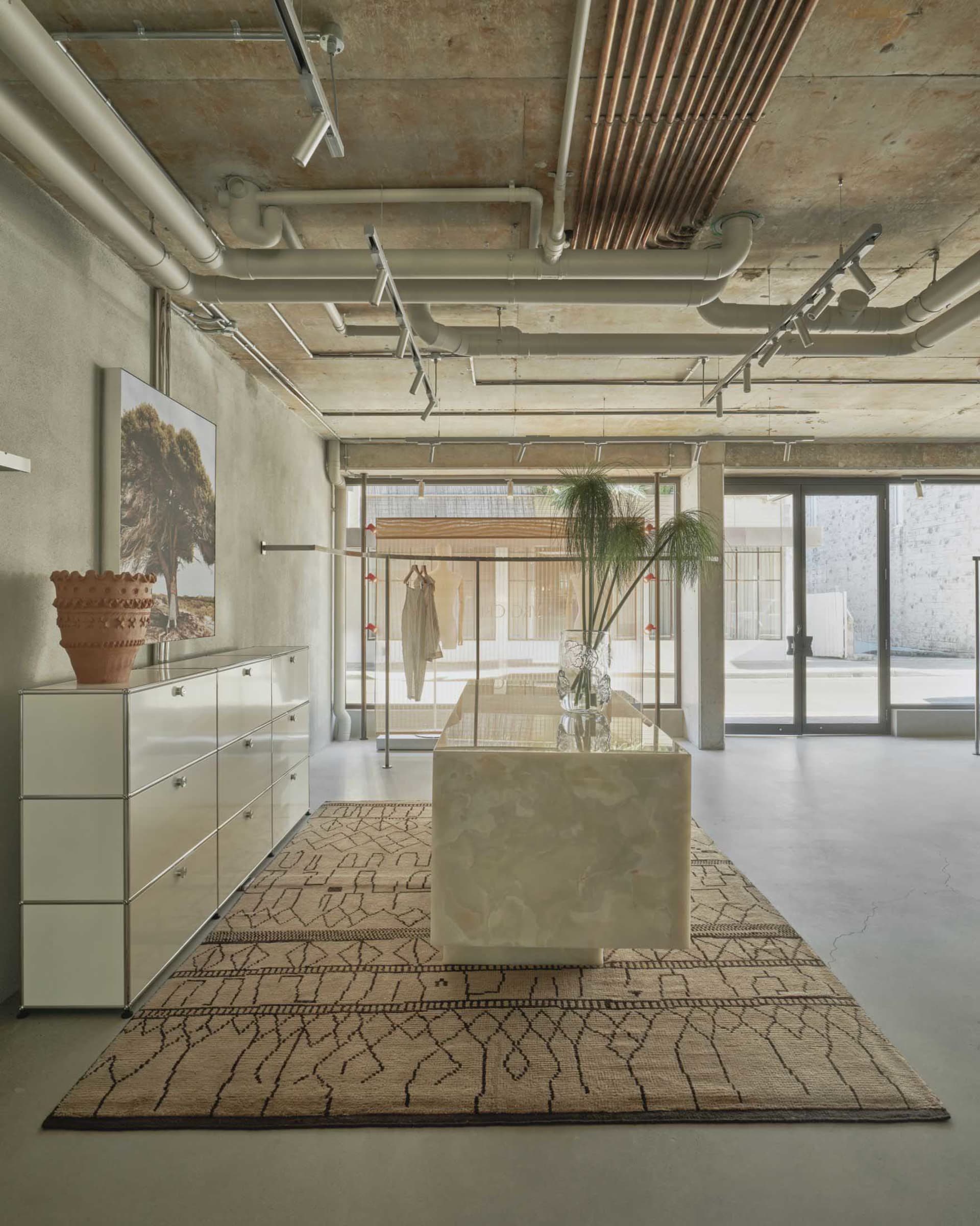 Modern minimalist boutique interior featuring a large onyx marble central island on a patterned rug, white modular cabinetry, concrete walls and ceiling with exposed white and copper pipes, and large glass doors at the back.