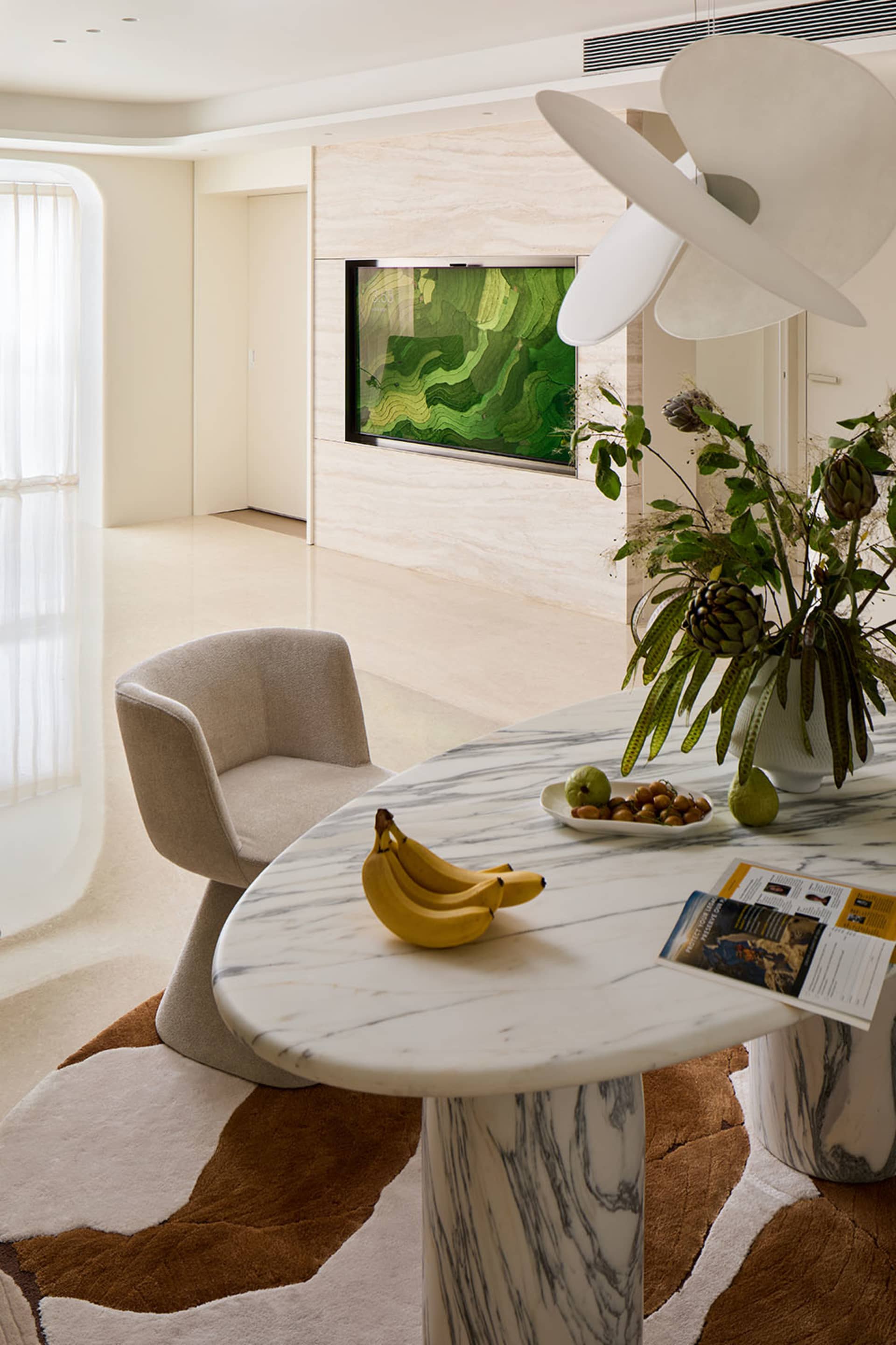 Modern dining area featuring a white and grey Calacatta marble oval table, a beige boucle swivel chair, a large abstract green artwork on a travertine wall, and a brown and white abstract rug.
