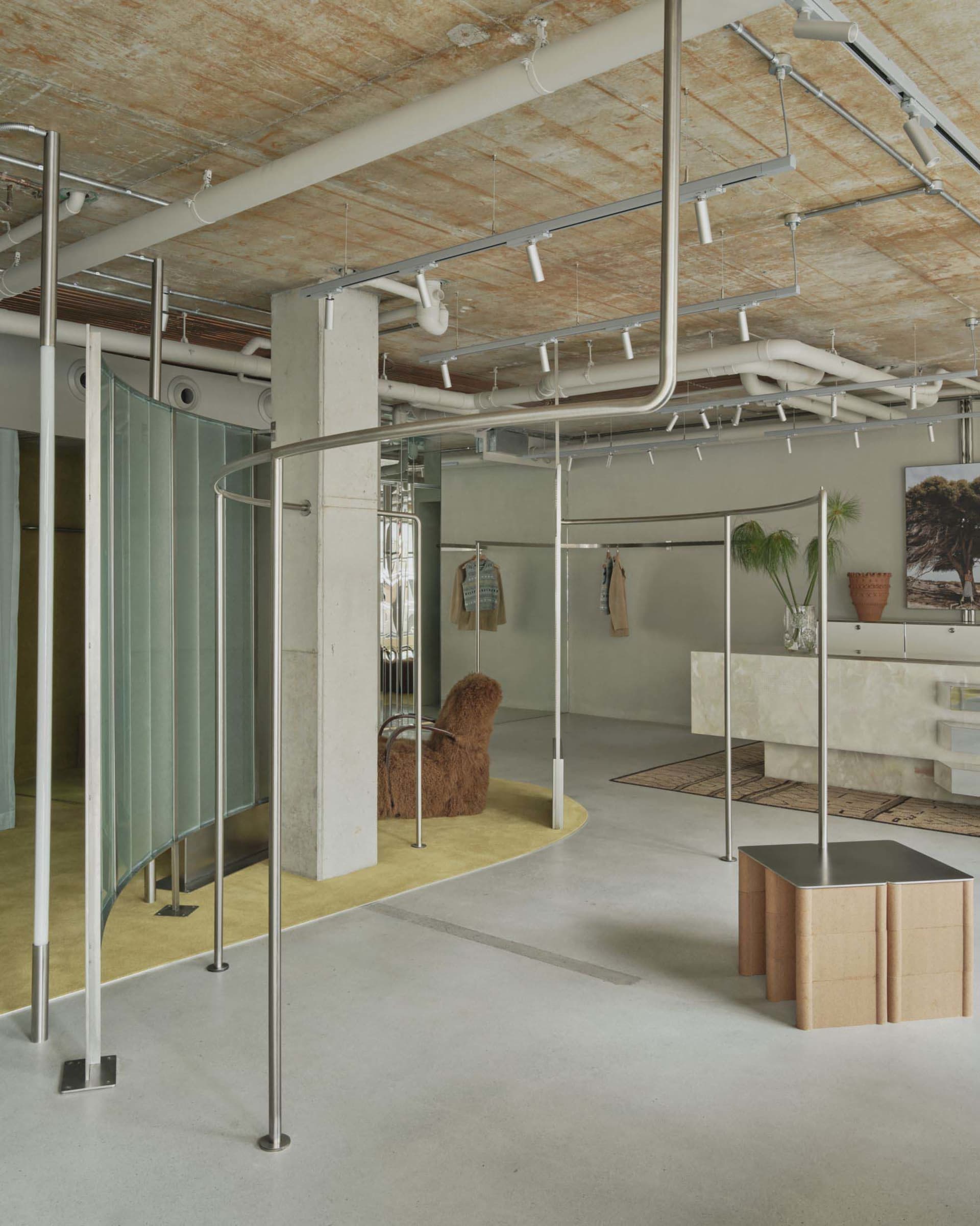 Modern boutique interior with curved stainless steel clothing display racks, polished concrete floor, exposed ceiling infrastructure, a shaggy brown armchair on a yellow rug, and a marble checkout counter.