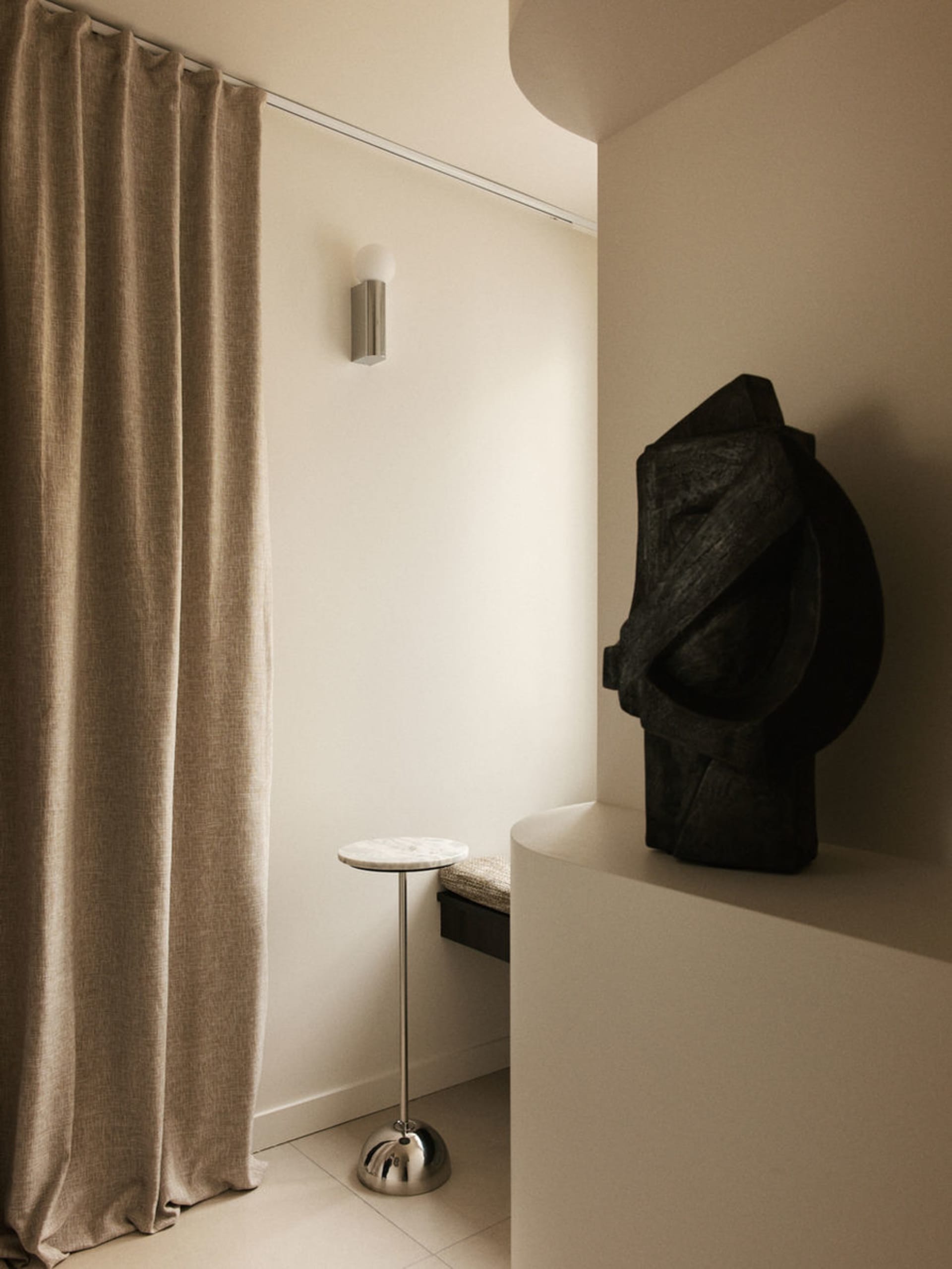 Modern minimalist entryway corner featuring a dark abstract stone sculpture on a curved white pedestal, a slender travertine-top side table with chrome base, beige linen curtain, and cylindrical wall sconce.