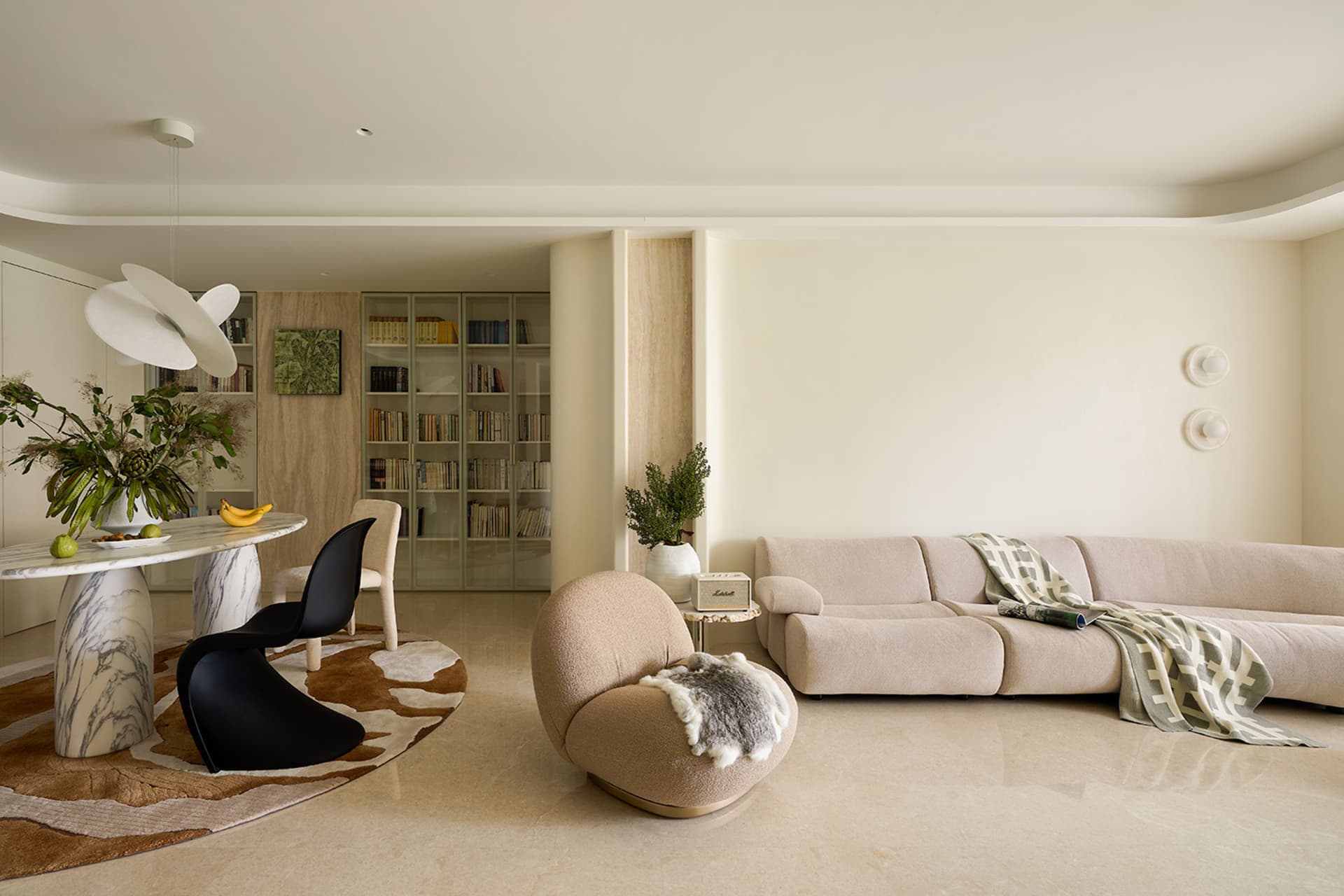 Modern minimalist living and dining area featuring a large beige sectional sofa, curved boucle swivel chair with sheepskin throw, marble dining table with black organic chair, built-in light wood bookshelf, and a cowhide rug on polished concrete floor.